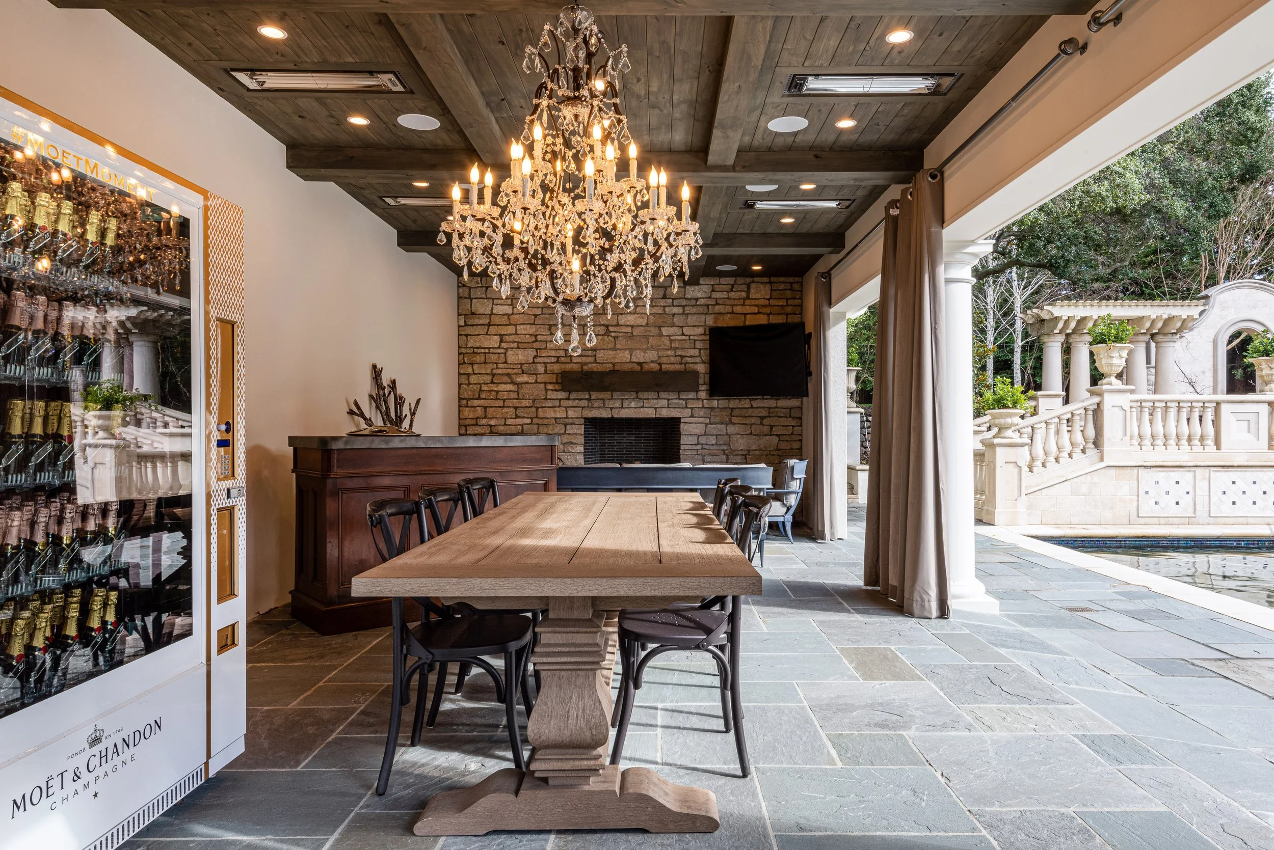 Indoor patio with a wooden dining table, black chairs, a chandelier, brick wall, fireplace, a flat-screen TV, and large open sliding doors leading to an outdoor patio with columns and a pool.