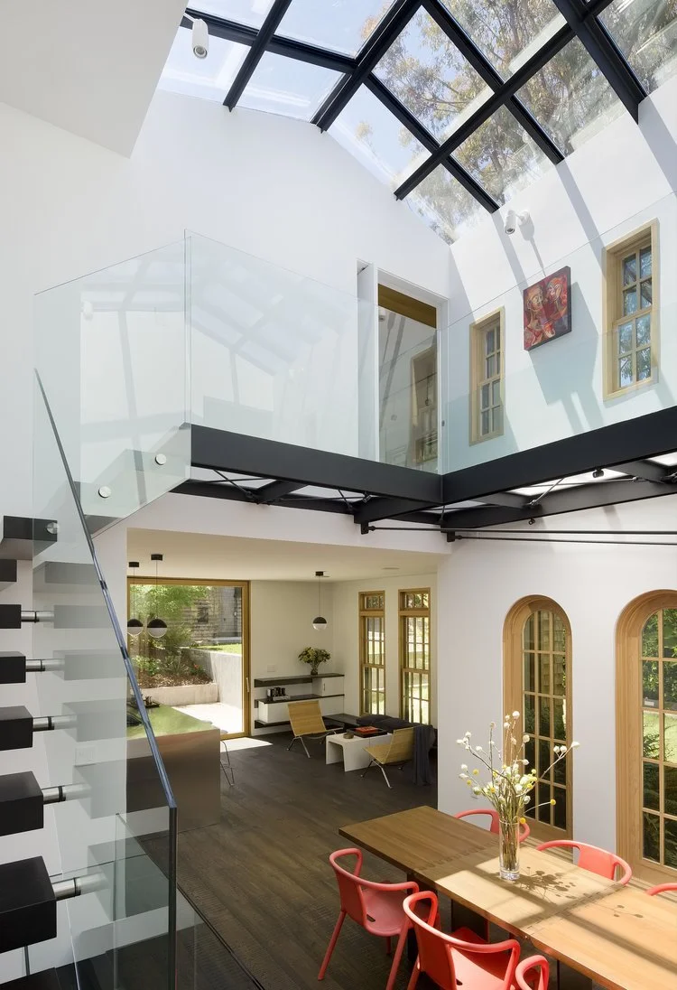 Interior of a modern, two-story house with a glass ceiling, white walls, and wooden accents. The ground floor has a dining table with pink chairs and a vase of flowers, a sitting area with armchairs and a window, and a staircase with black steps and 