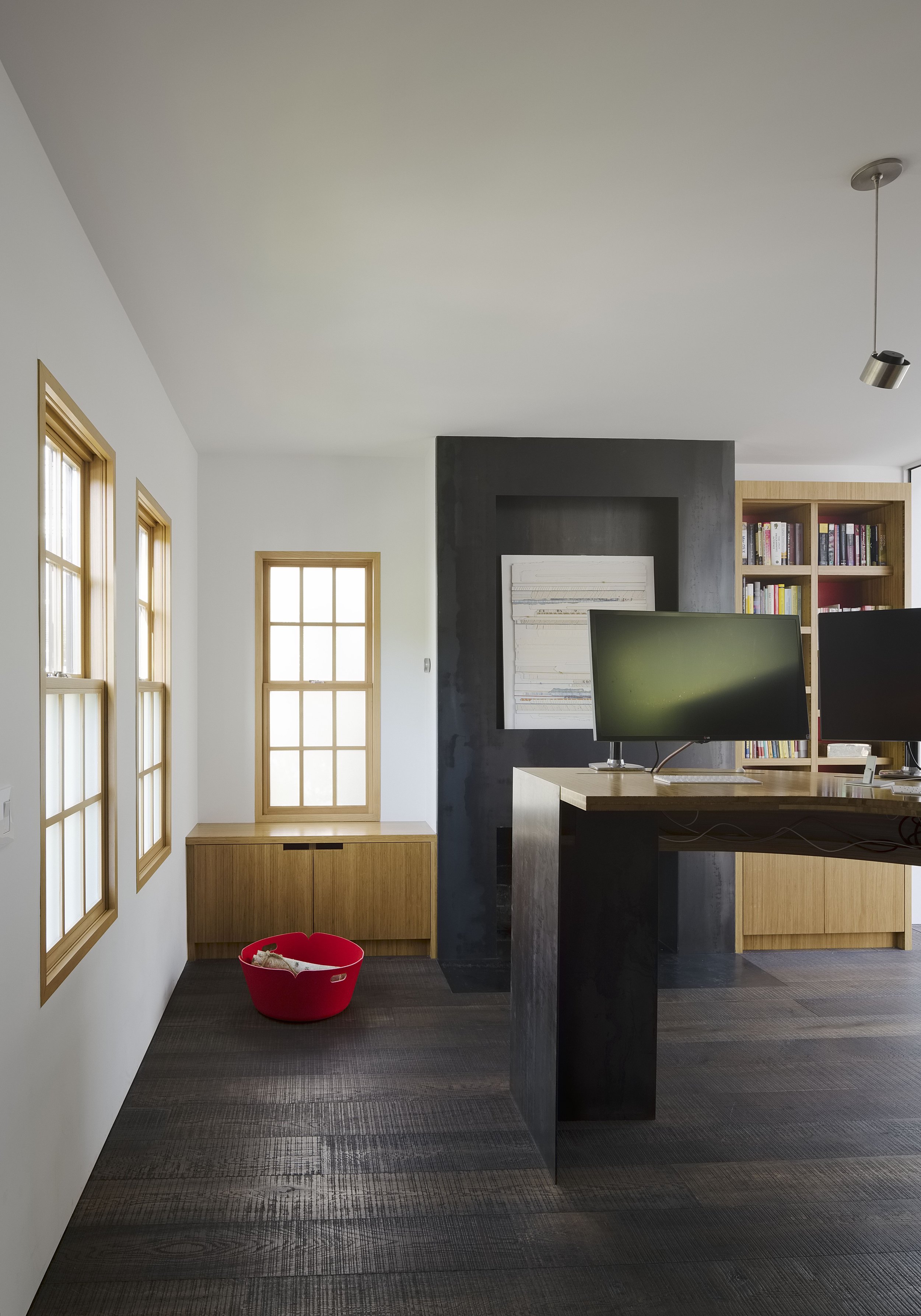 Modern office space with three windows on the left wall, a small cabinet below the middle window, a red pet bed on dark wooden floor, black wall with white framed art, and wooden bookcases with colored books. Two computer monitors on a wooden desk ar