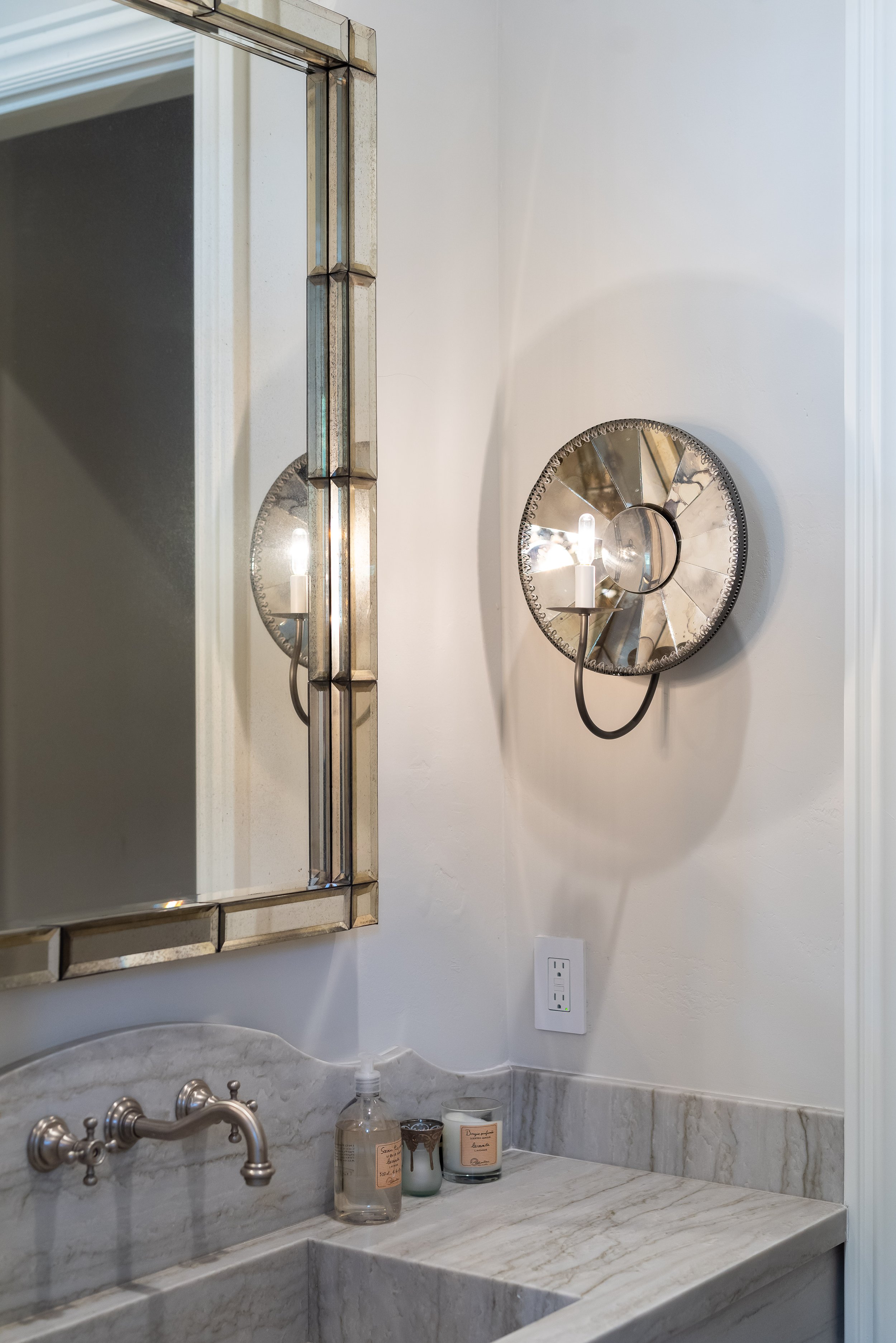 Bathroom with marble countertop, a mirror with a decorative frame, a wall-mounted light fixture with a reflective silver backing, and candles and soap on the countertop.