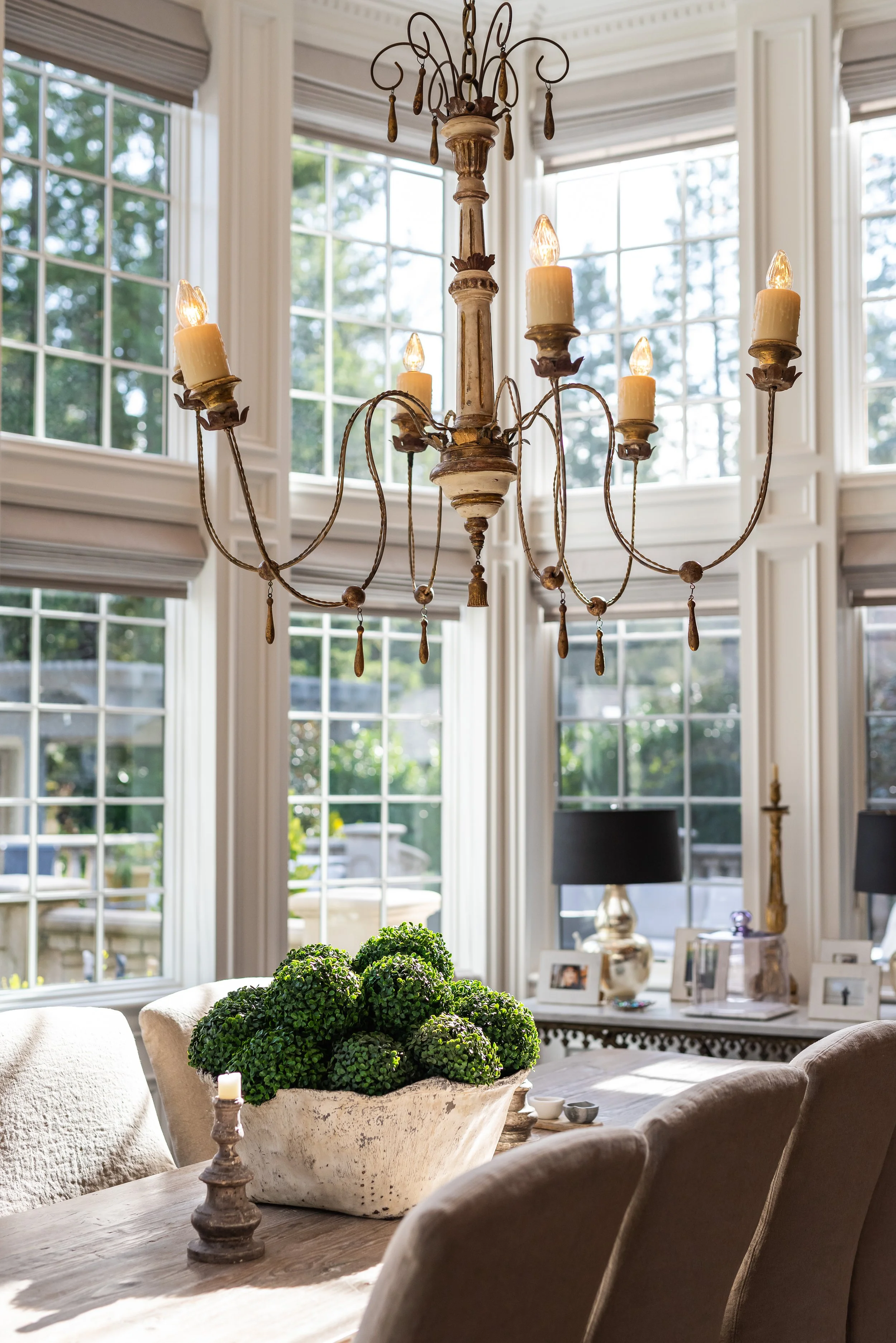 Elegant dining room with a rustic chandelier, large windows with blinds, a table with a large white pot of green plants, and framed photos with lamps in the background.