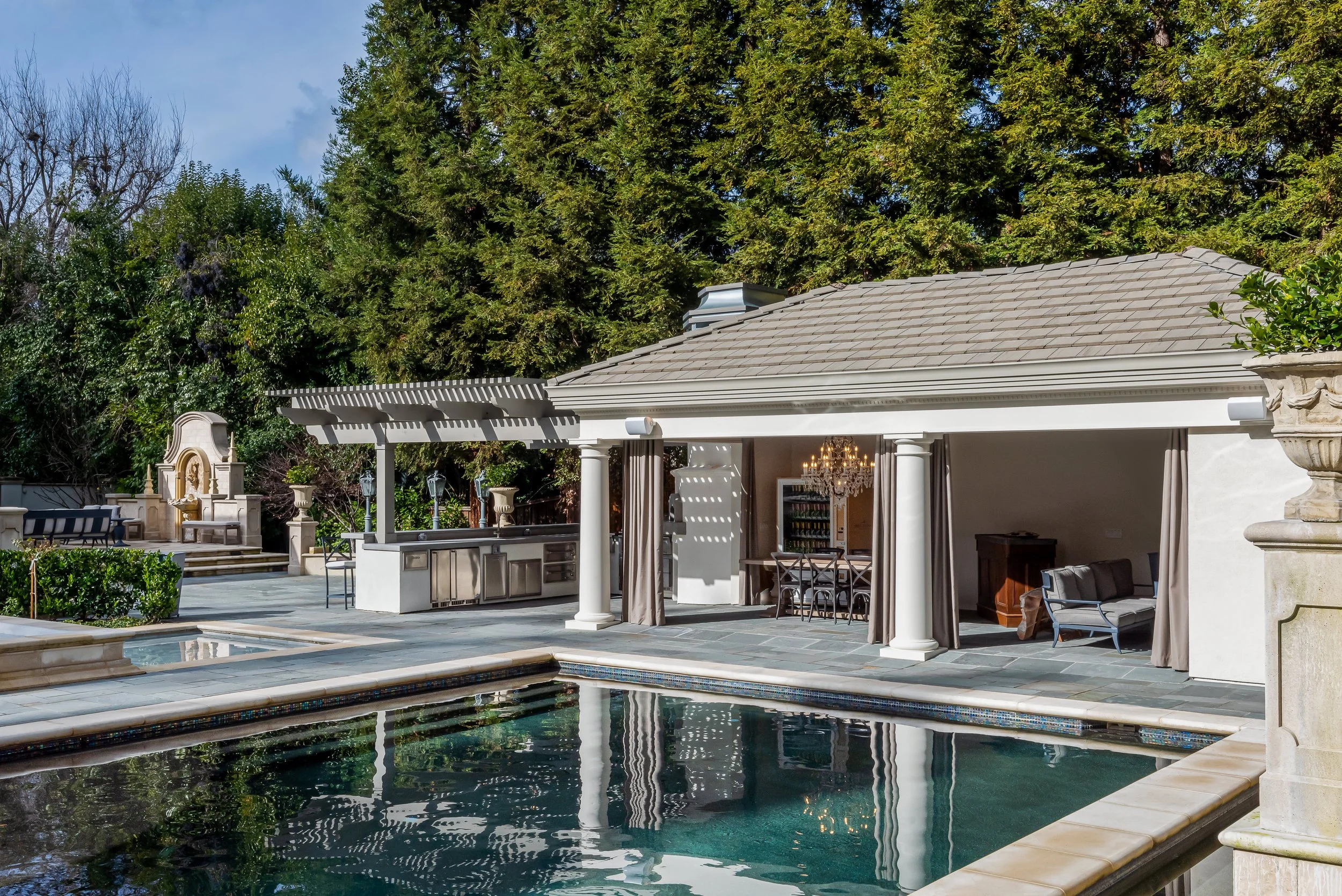 Luxury backyard with a swimming pool, shaded outdoor bar area with curtains, patio furniture, a chandelier, and lush trees in the background.