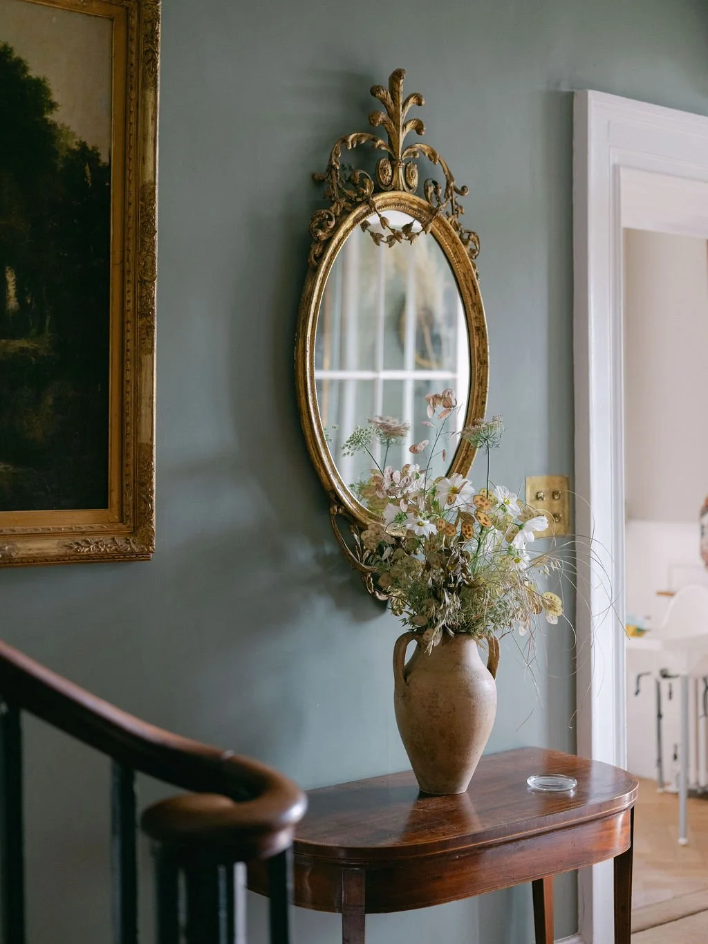 An oval gilded mirror with ornate scrolling details hangs on a light blue wall. Below the mirror, a wooden table holds a large ceramic vase filled with dried flowers and greenery.