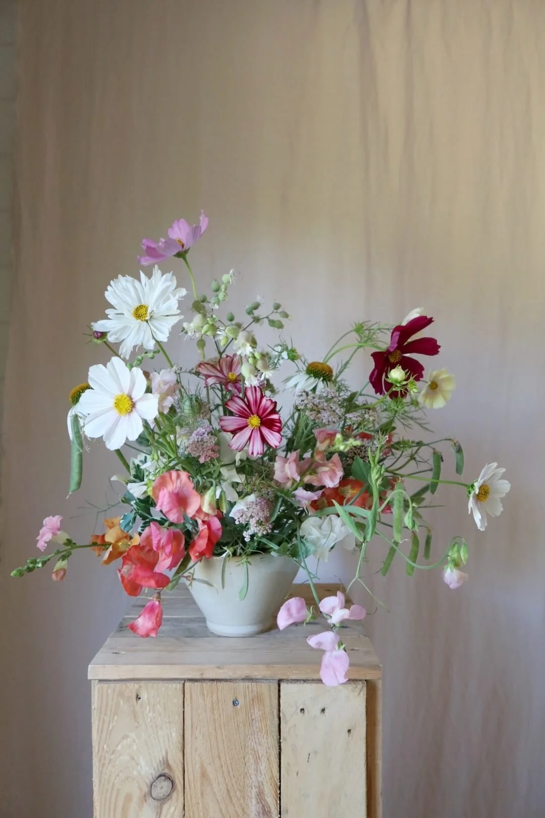 A colorful bouquet of various flowers in a white vase on a wooden pedestal against a neutral background.