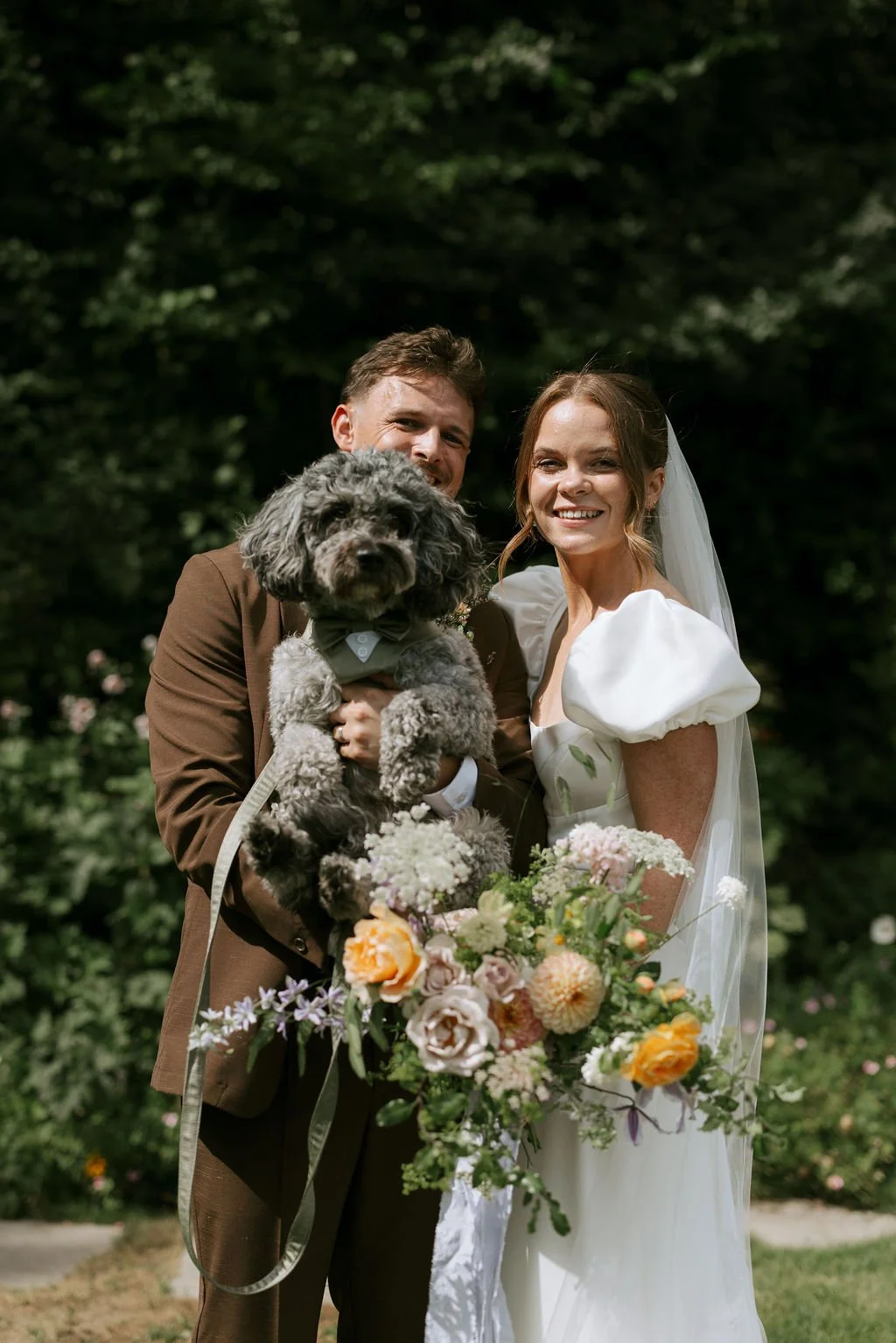 A smiling bride and groom holding a fluffy dog in a garden with greenery behind them. The bride is holding a colorful bouquet of flowers.