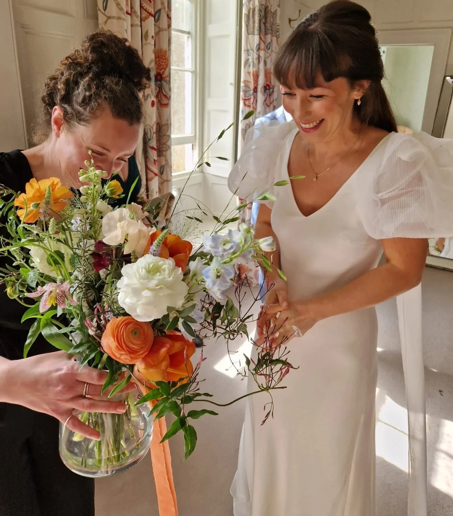 Two women, one a bride in a white dress and another in black the wedding florist, look at a large coloruful flower bouquet in a clear glass vase, laughing and smiling in a bright room with floral curtains.