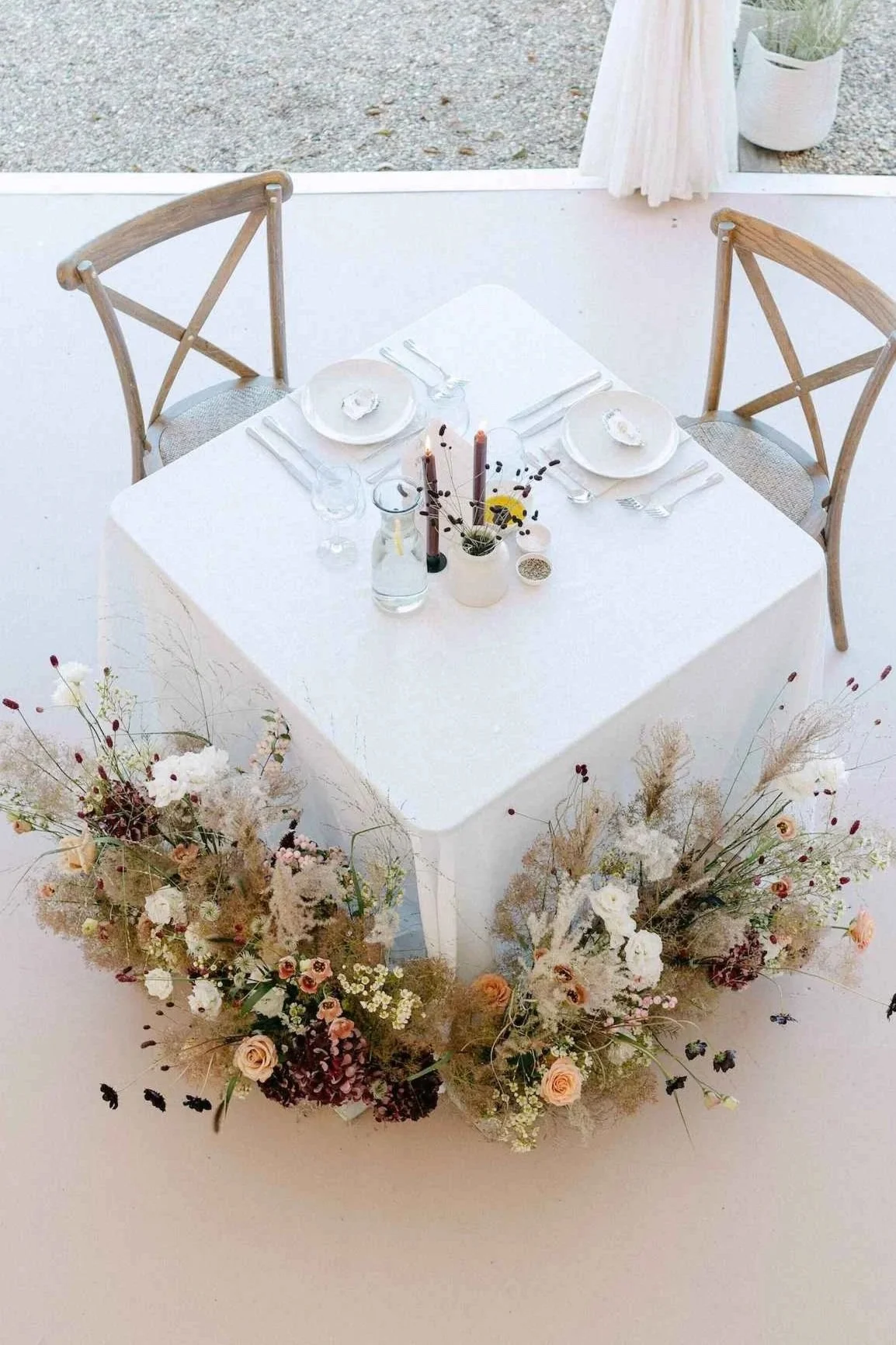 A white table with two wooden chairs, set for a meal with plates, cutlery, and candles, decorated with floral arrangements on the floor.