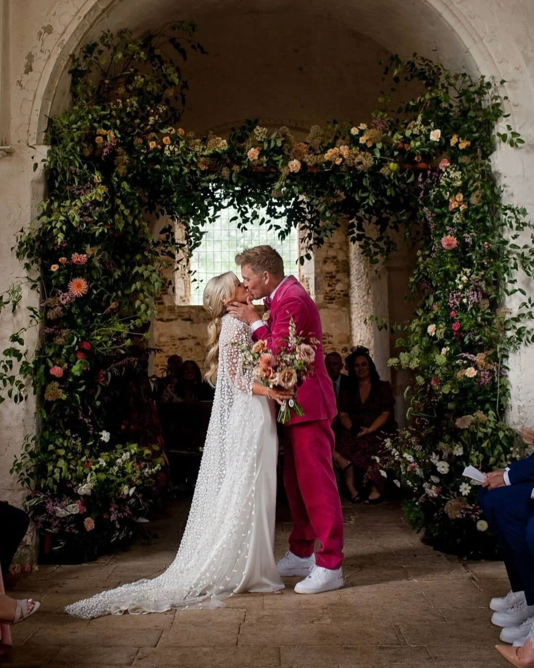 A bride and groom at their wedding kiss under a floral arch inside a rustic stone building, with guests seated around them.