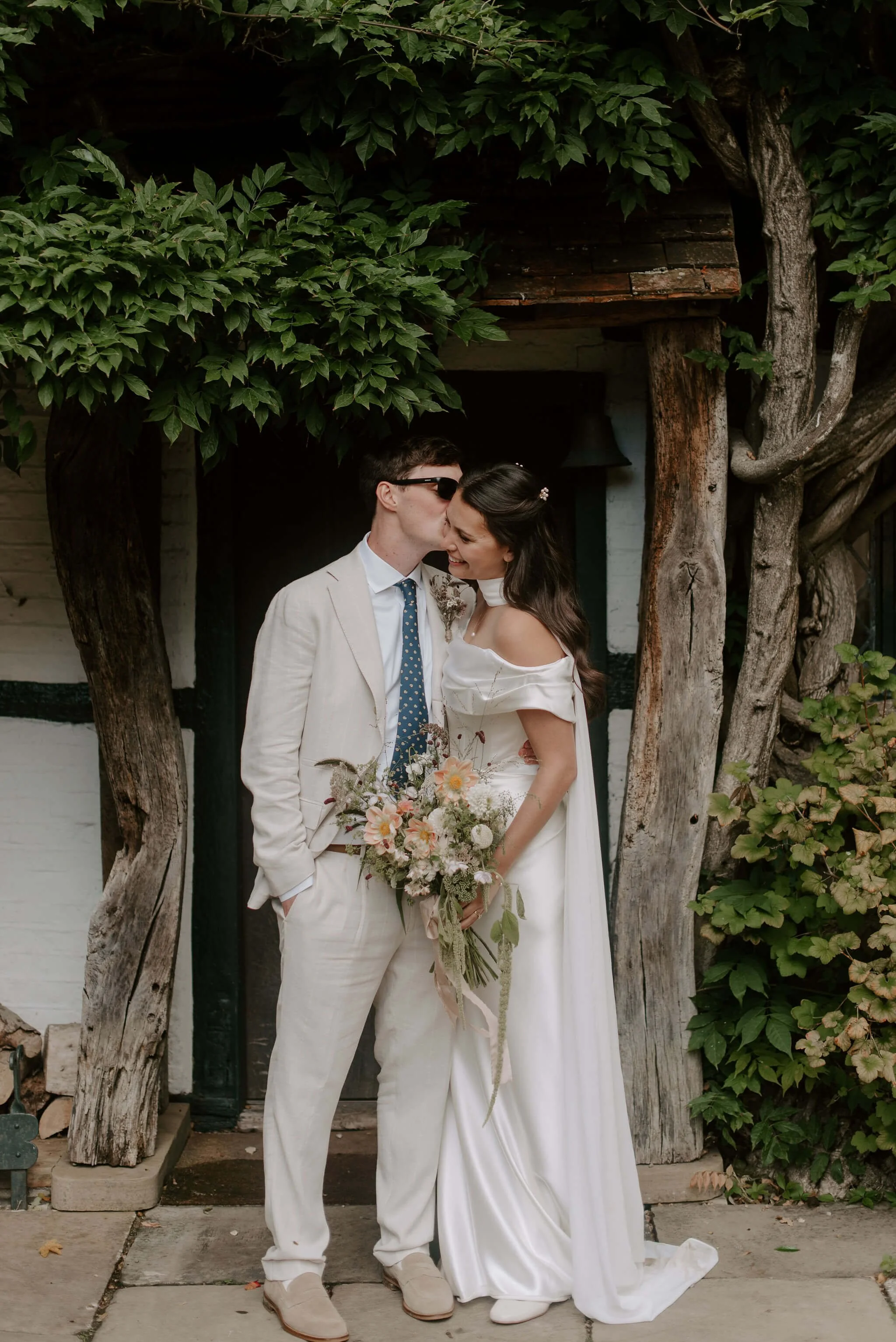A bride and groom sharing a kiss in front of a rustic wooden structure surrounded by green foliage. The bride wears a white off-shoulder wedding dress and holds a bouquet, while the groom is dressed in a light-colored suit with a blue polka dot tie, sunglasses, and has his hands in his pockets.