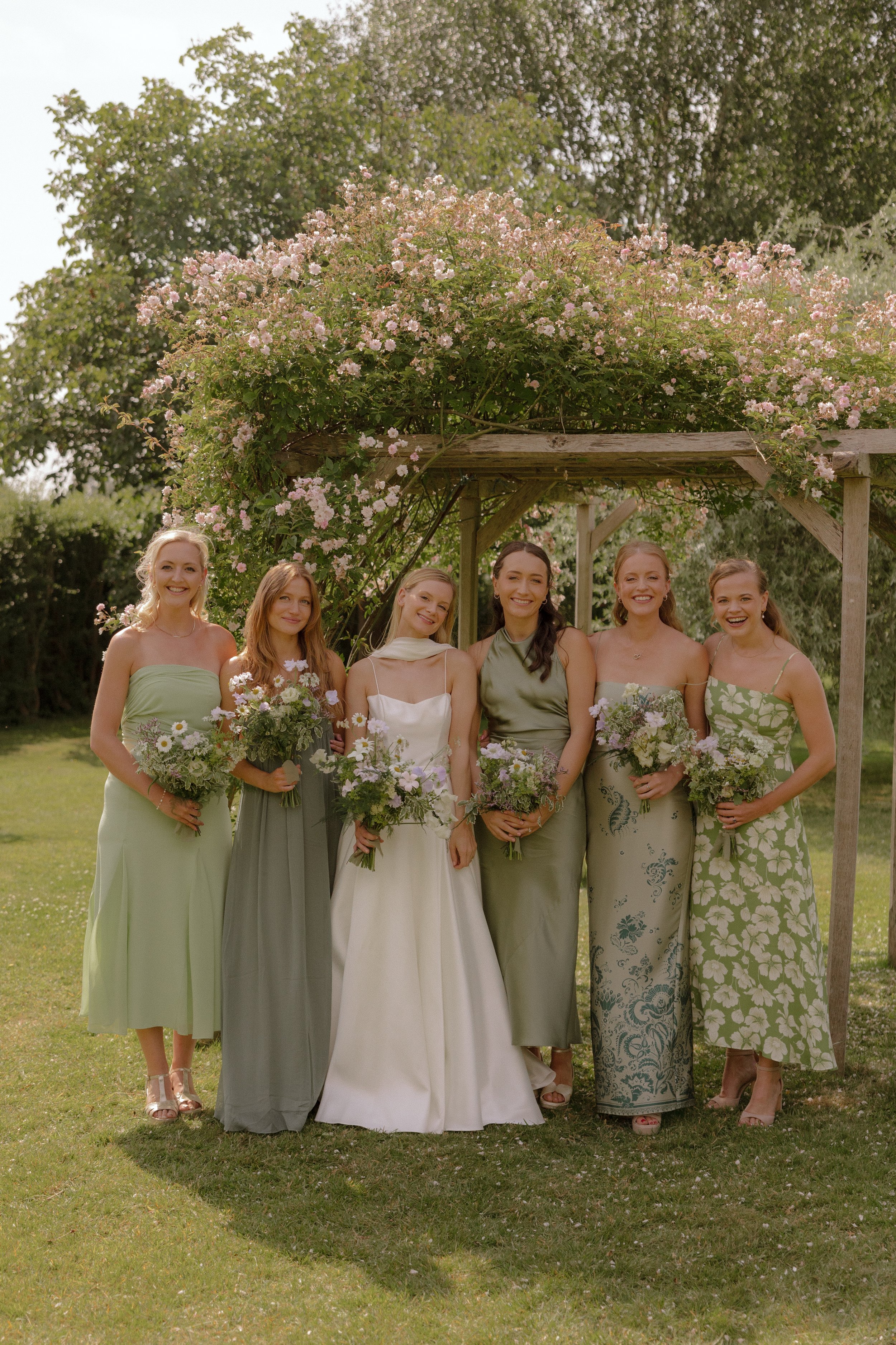 Group of six women in pastel dresses holding bouquets of flowers, standing under a wooden arch covered in pink and white flowers on a grassy lawn during daytime.