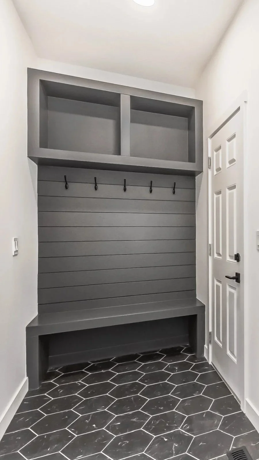 Empty mudroom with white walls, a gray built-in organization unit with hooks and storage shelves, black hexagonal tile flooring, and a white door.