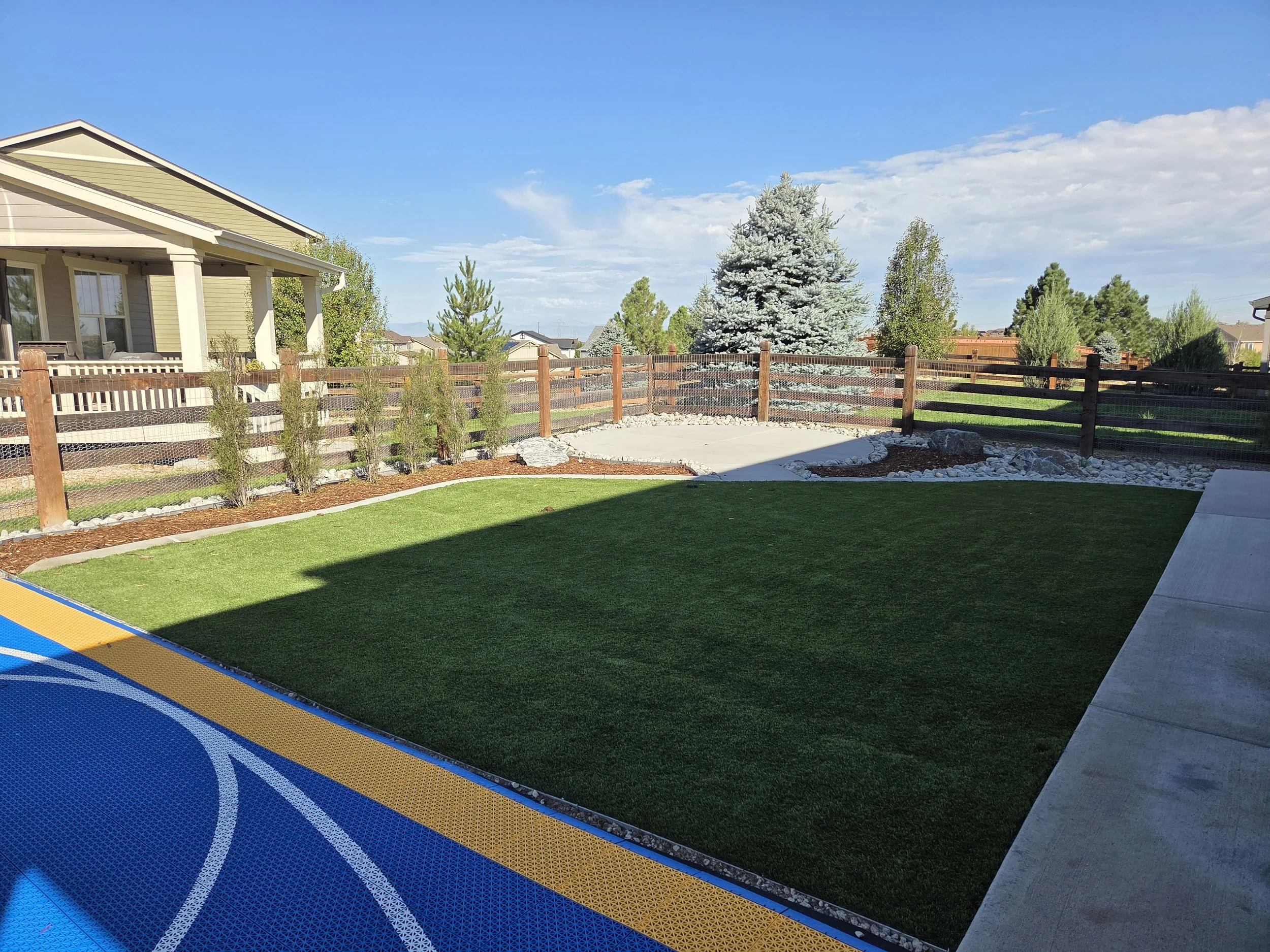 A backyard with a porch, a large evergreen tree, and a wooden fence under a blue sky with clouds.
