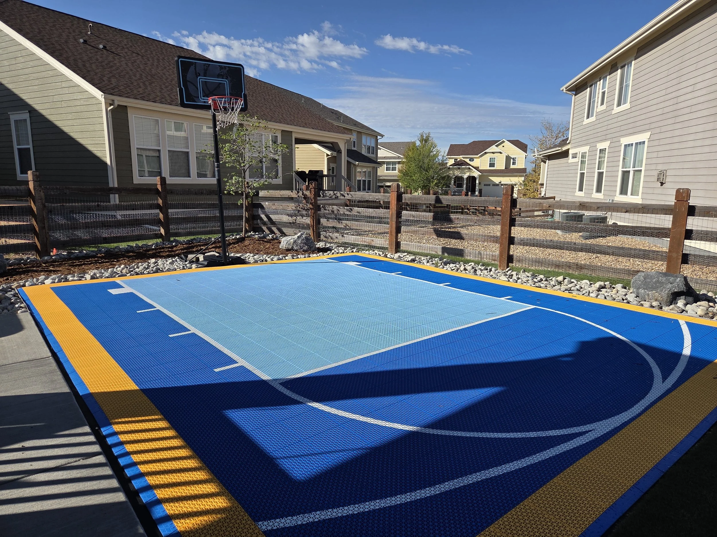 Backyard with a portable basketball hoop and a blue, orange, and white mini basketball court, surrounded by a wooden fence and neighboring houses under a partly cloudy sky.