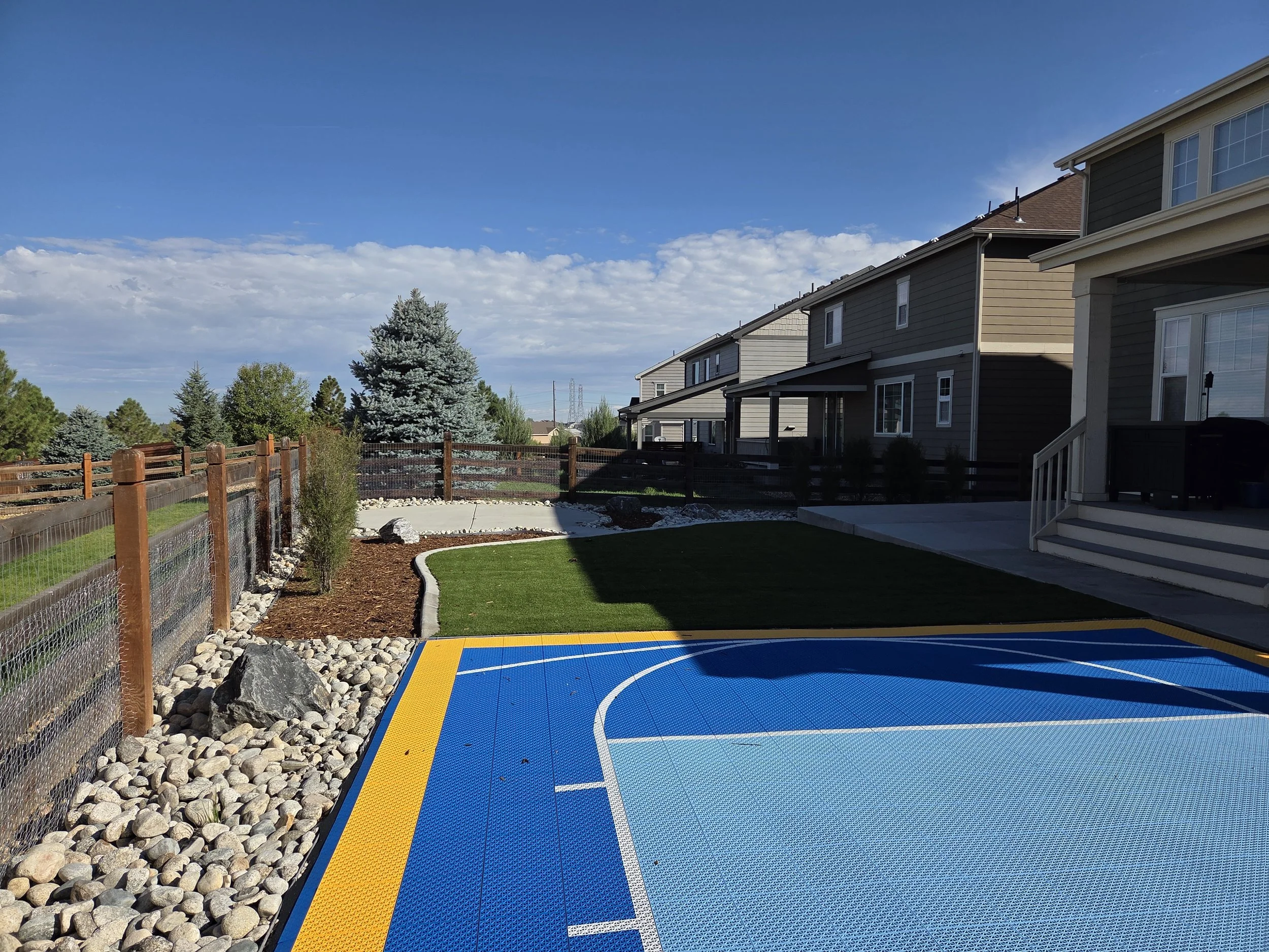 Backyard with playground surface, artificial grass, large rocks, wooden fence, trees, and modern houses under a partly cloudy sky.