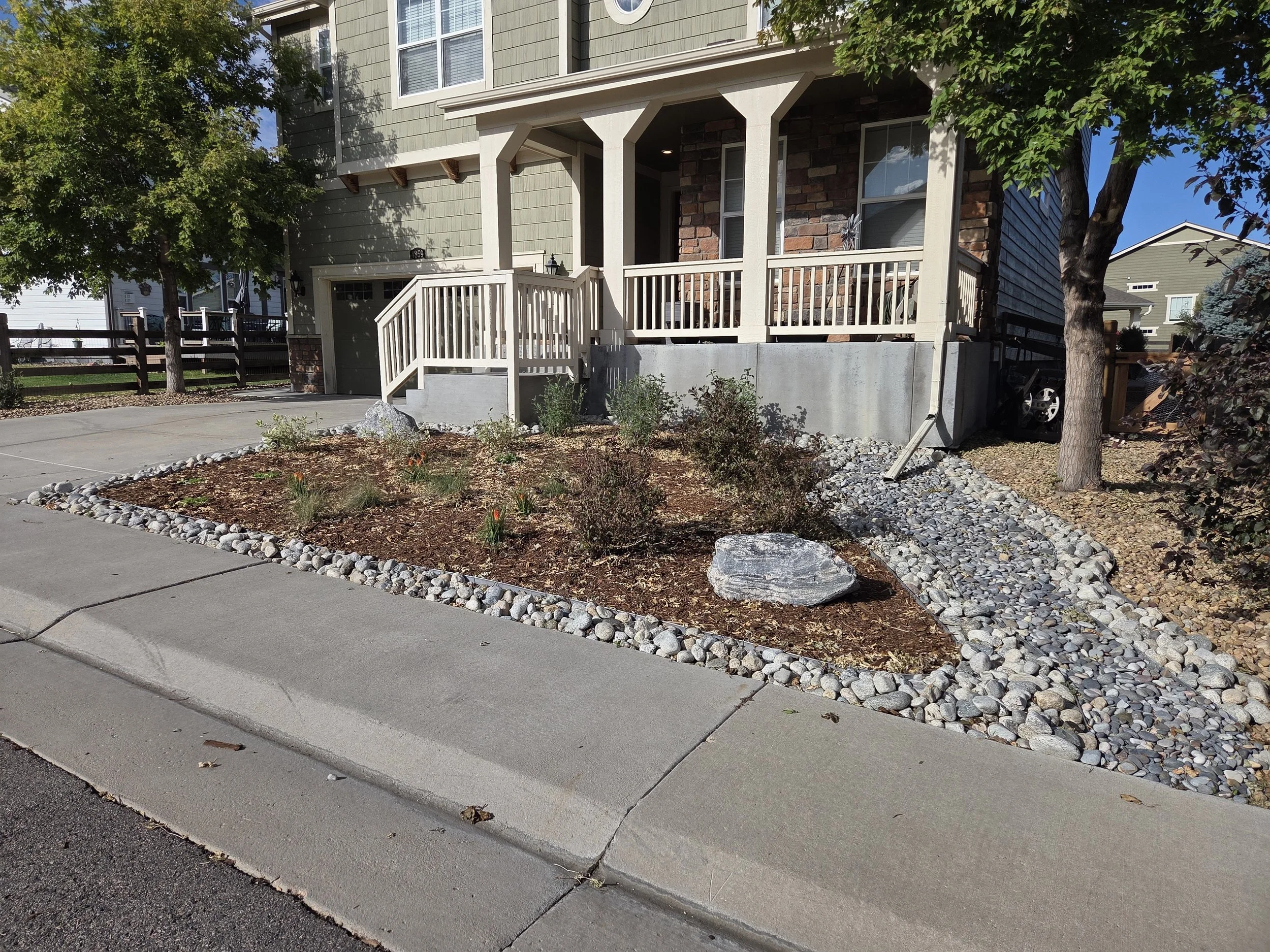 Front yard of a house with a small porch, stairs, and a garden bed bordered by rocks. A tree is on the right side, and other houses are visible in the background.