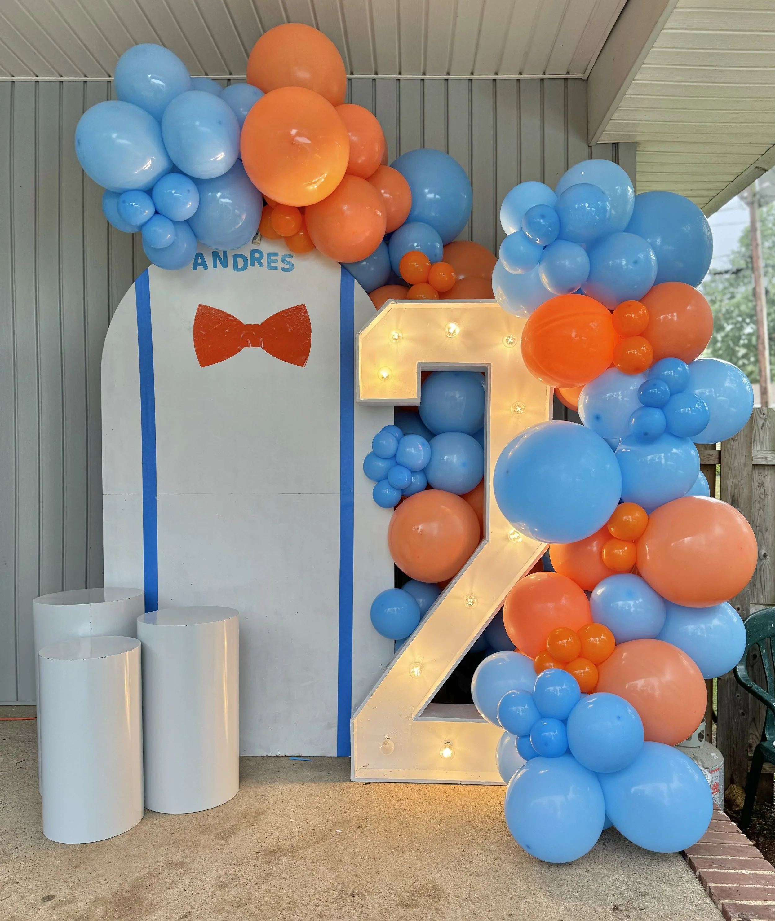 Decorative birthday display with a large illuminated number 2 surrounded by blue and orange balloons, a whiteboard with the name Andres and a red bow tie, three white cylindrical pedestals, set against a gray paneled wall.