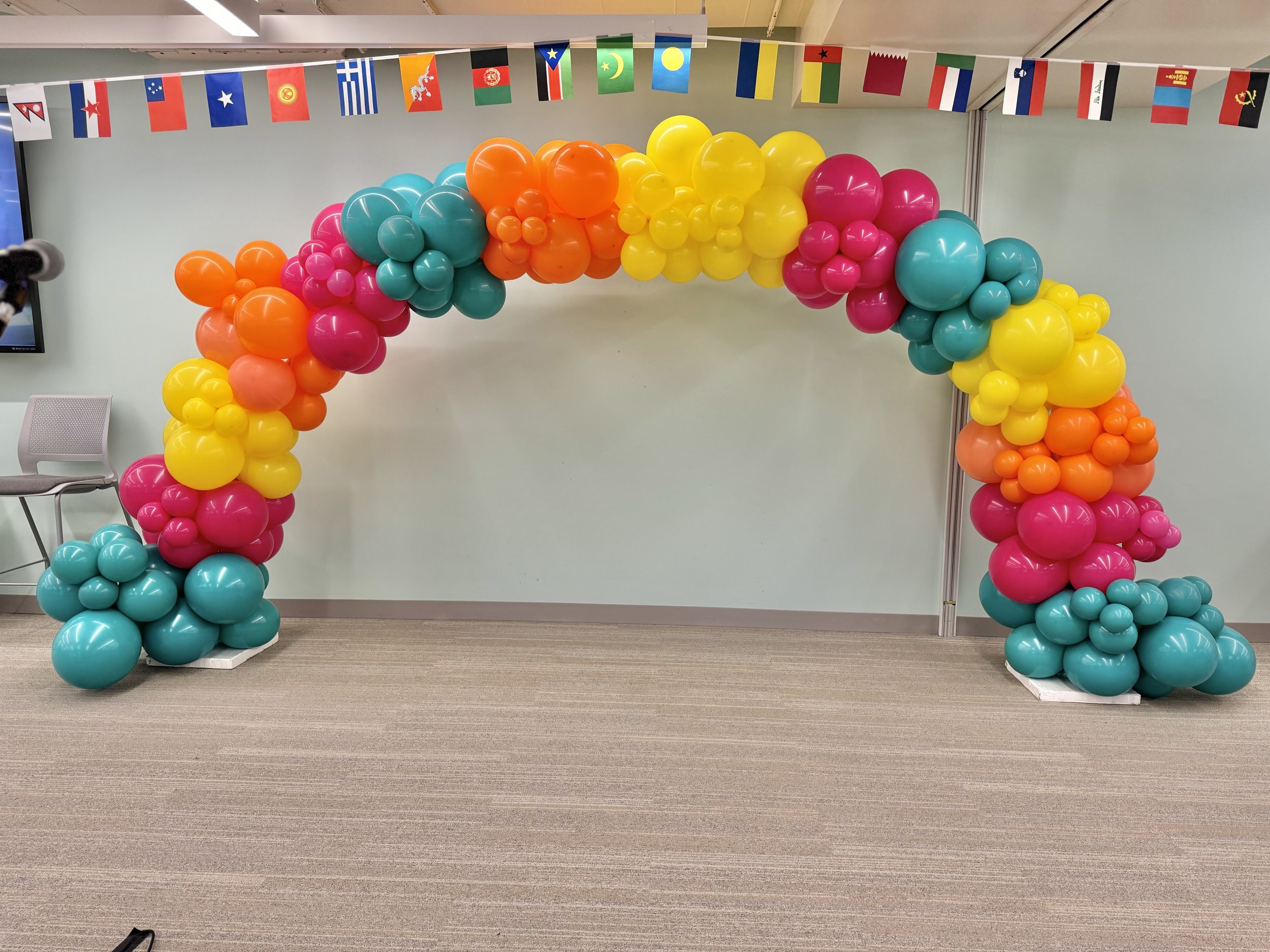 Colorful balloon arch with pink, yellow, orange, and teal balloons, decorated with international flags hanging above, placed in an indoor setting.