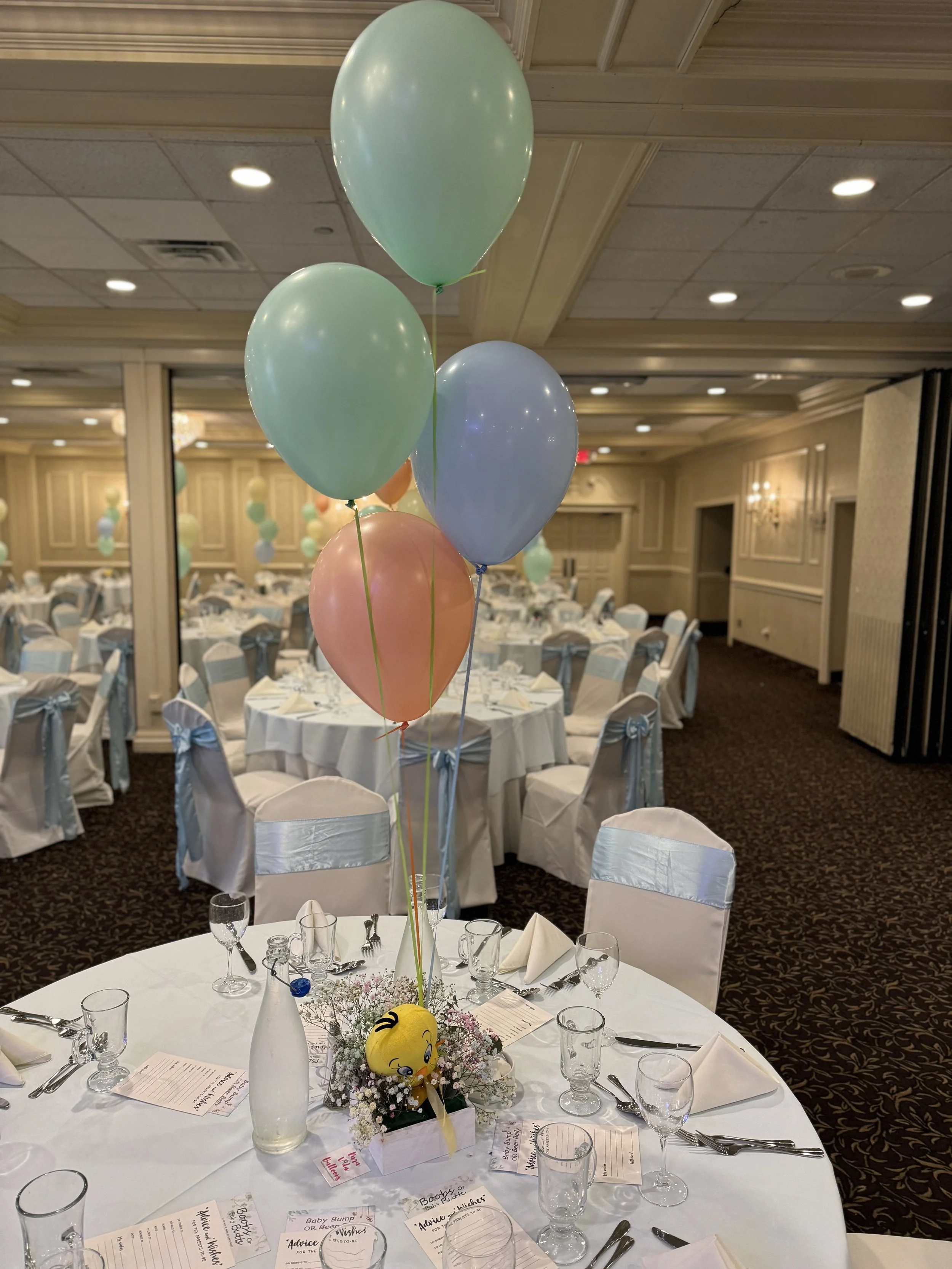 Round banquet table decorated with a centerpiece of pastel-colored balloons, a small yellow emoji plush toy, and flowers, set in a decorated event hall with white tablecloths, folded napkins, and set glassware.