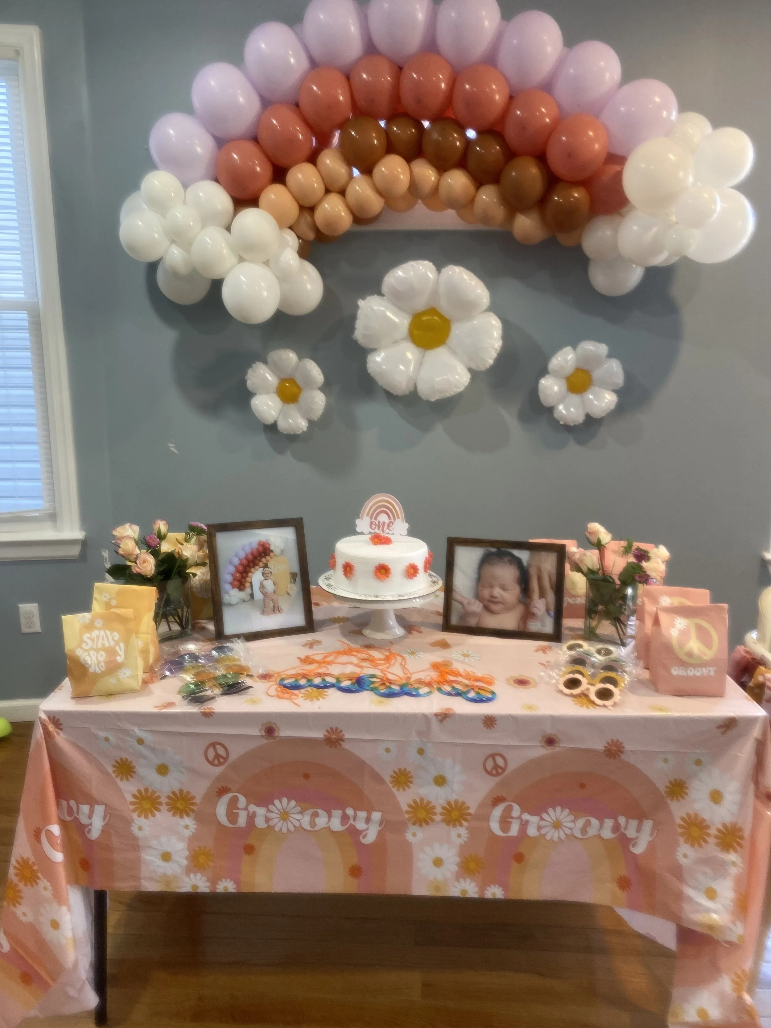 Table decorated for a first birthday with a colorful rainbow balloon arch, framed pictures of a baby, a cake with a rainbow and 'one' topper, and flower and peace sign decorations.