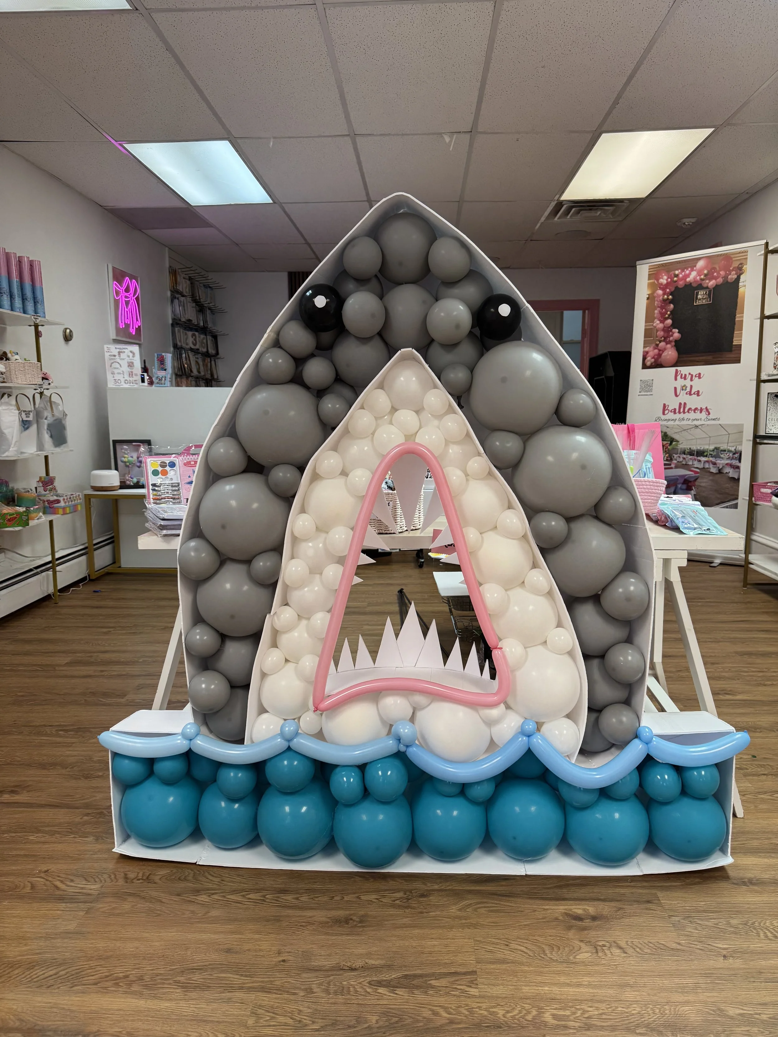 Balloon decoration resembling a birthday cake or dessert with layers of blue, gray, and white balloons, topped with a pink balloon outline of a triangle or open mouth with sharp points, set up at a party or store display.