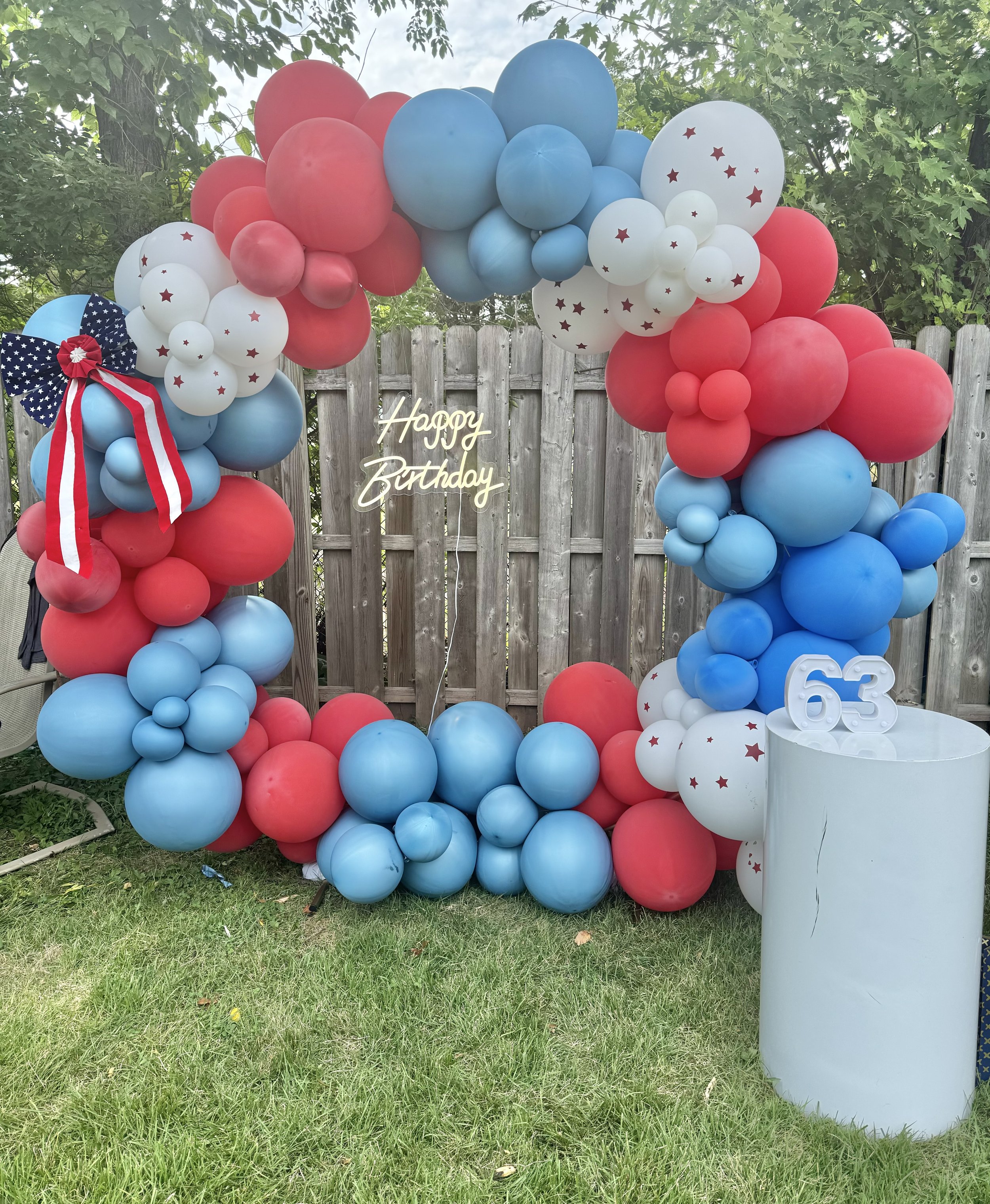 Red, white, and blue balloon arch with a neon 'Happy Birthday' sign, a white cylinder with the number 63, and a wooden fence with green trees in the background.