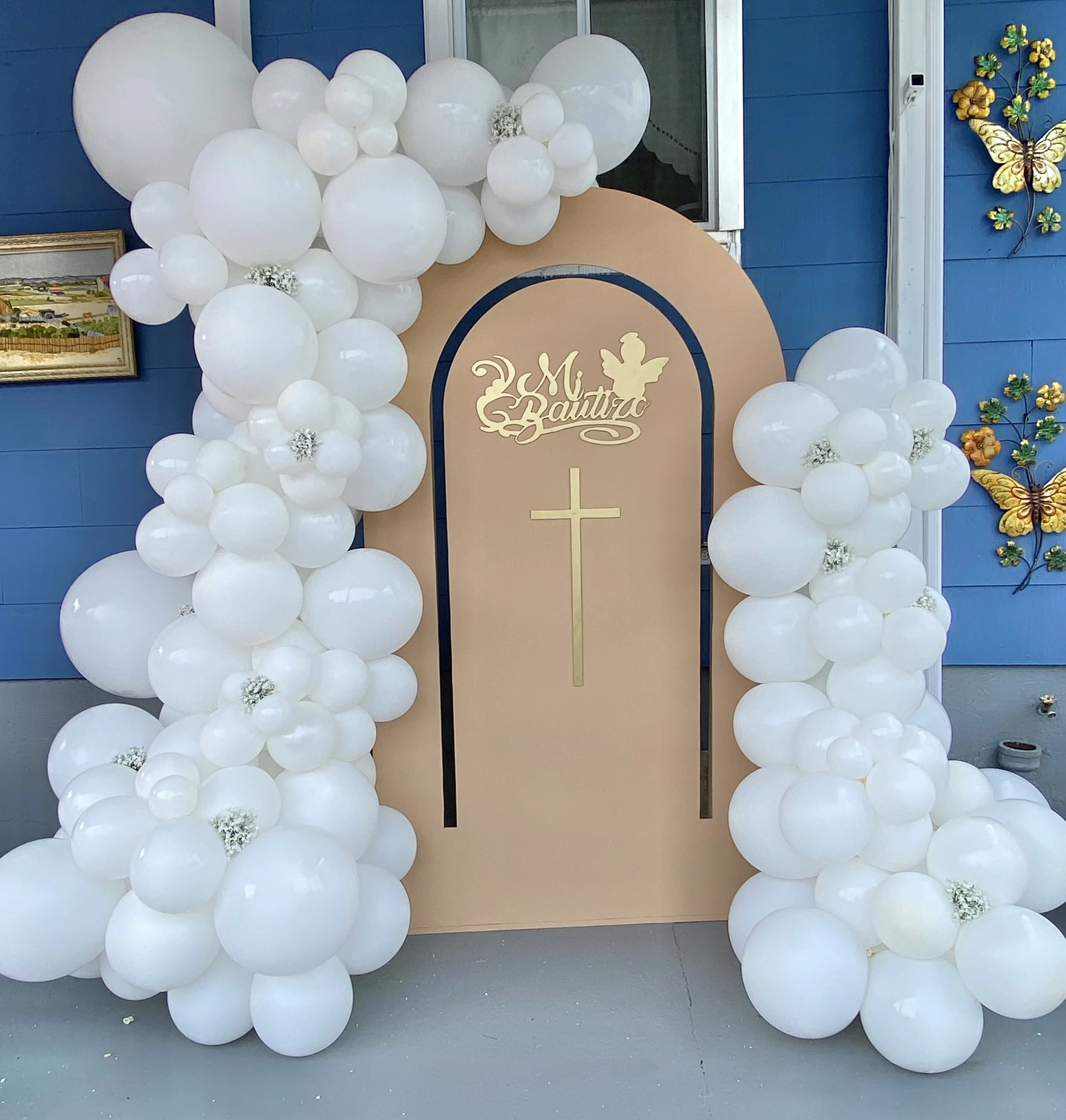 Decorative setup with white balloons forming an arch around a beige and dark blue backdrop. The backdrop features a gold cross and a script that appears to read 'Мира' with a small angel silhouette. There are colorful butterfly wall decorations on th
