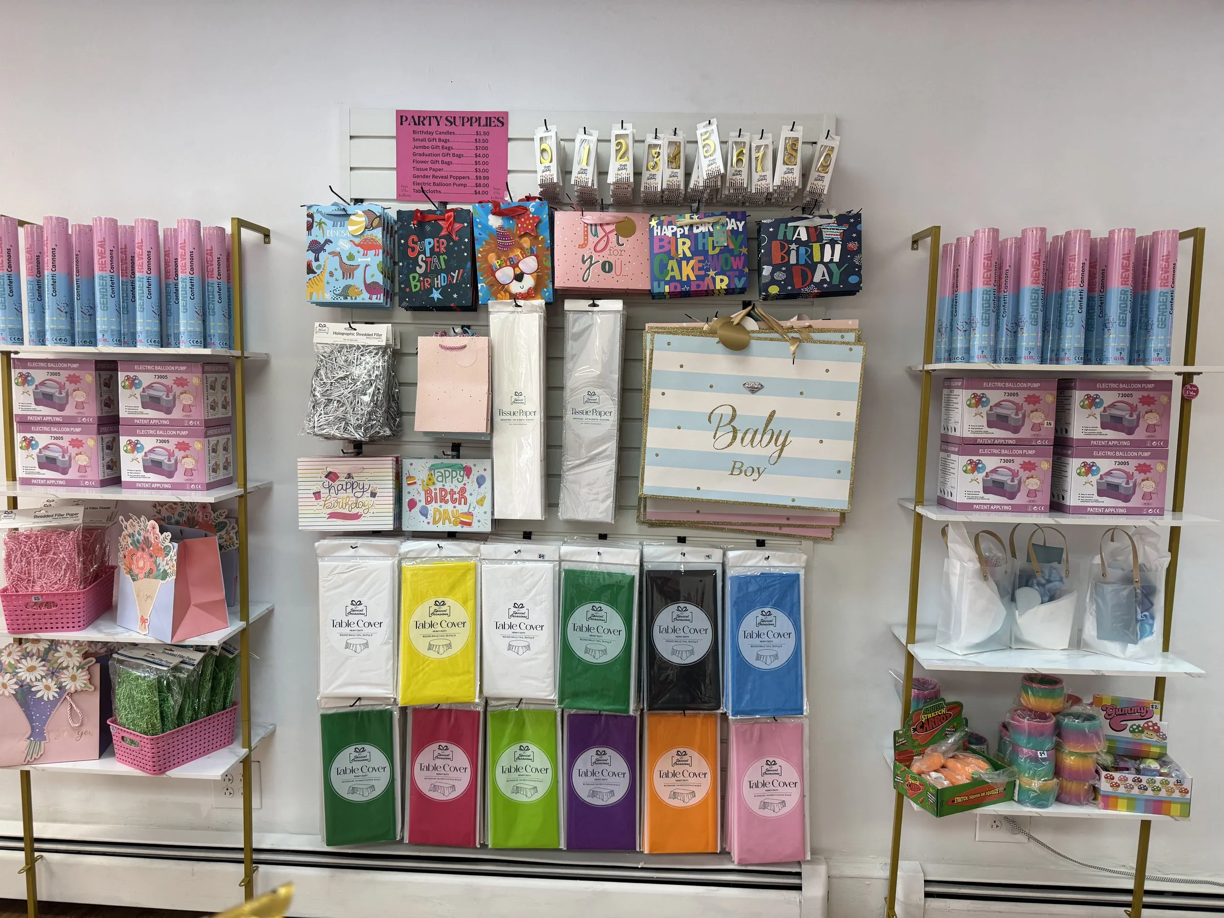 A store shelf displaying party supplies including gift bags with birthday and other themes, tissue paper in various colors, and table covers. Signage indicates pricing for party-related items.