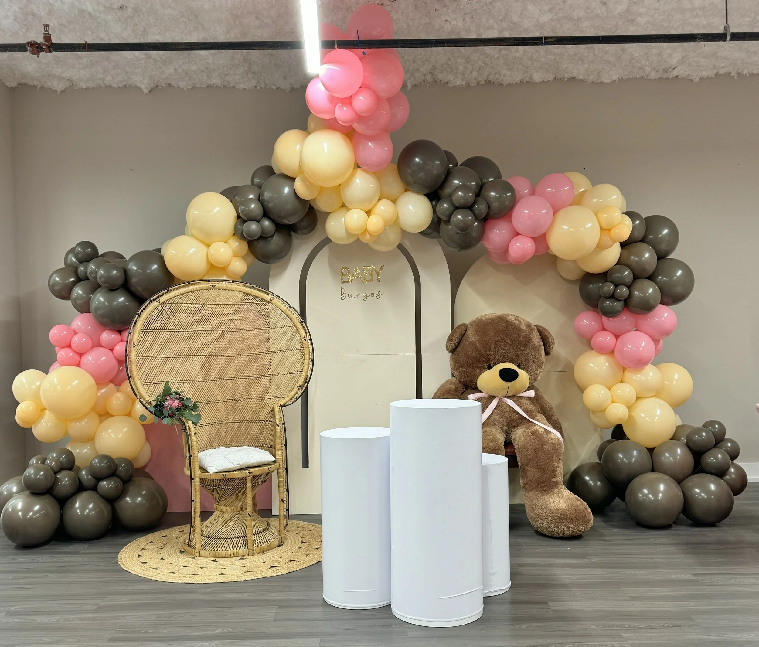 Decorative setup with a balloon arch in pink, beige, and gray, a wicker peacock chair with a floral arrangement, white pedestals, a large teddy bear, and a backdrop with the words "BABY Burgos" for a baby shower.