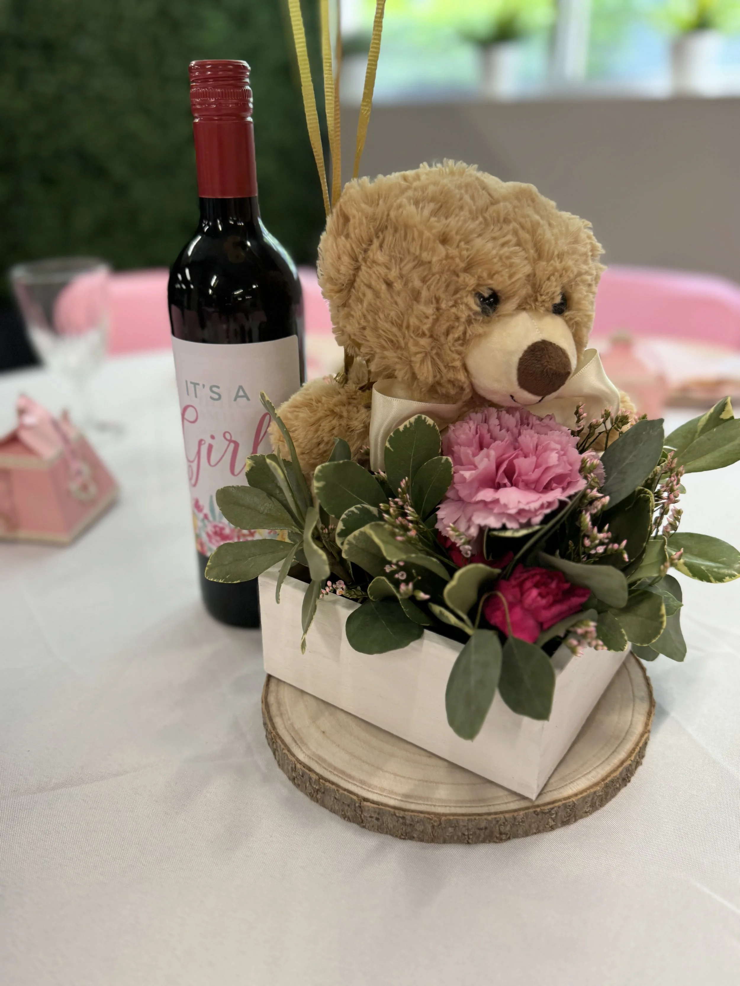 Decorative arrangement with a plush teddy bear, pink and green flowers in a white box on a wood slice, with a bottle of red wine labeled "It's a Girl" in the background.