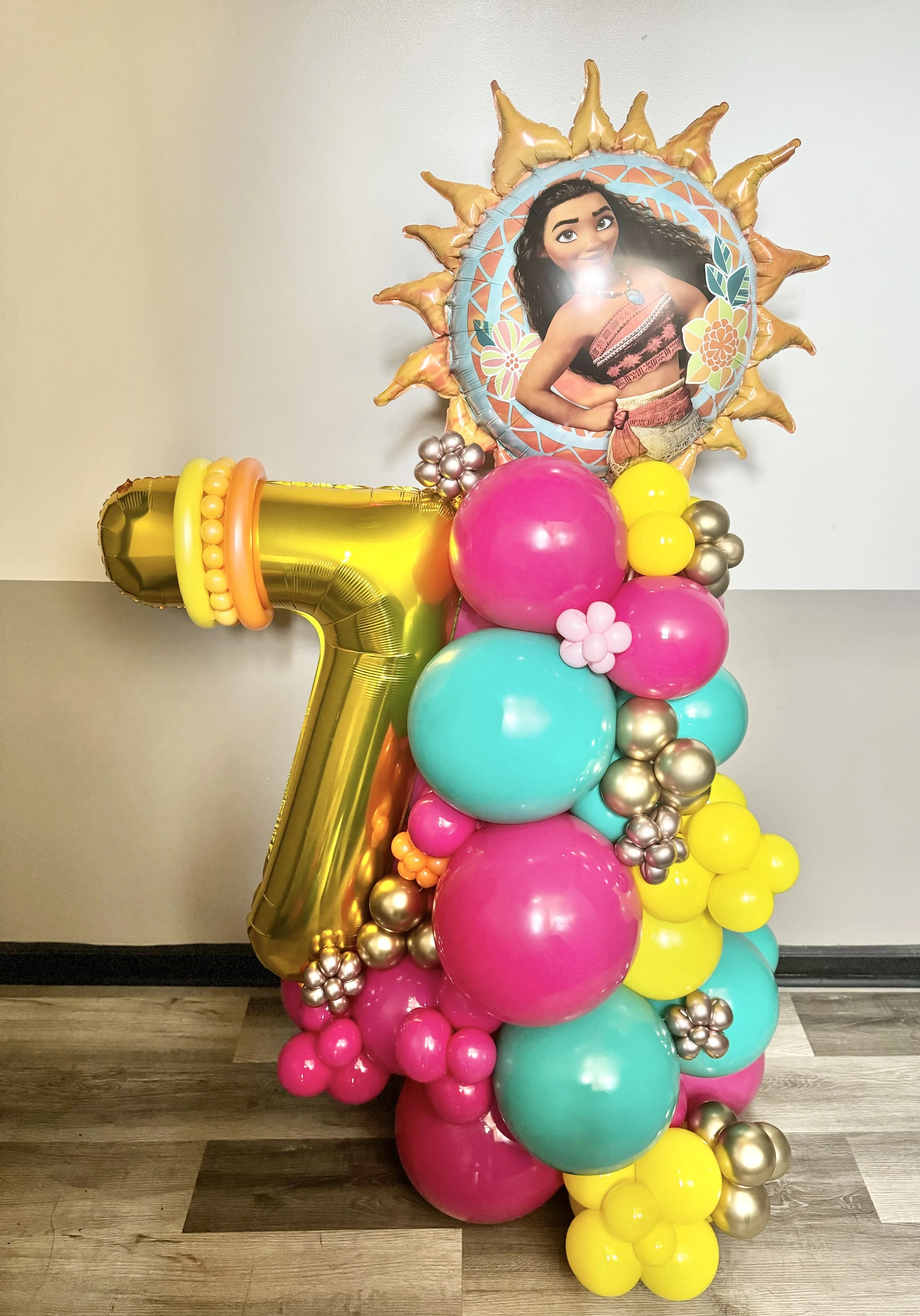 Colorful balloon arrangement featuring a large gold number seven balloon, a Moana character balloon, and clusters of pink, blue, yellow, and silver balloons.