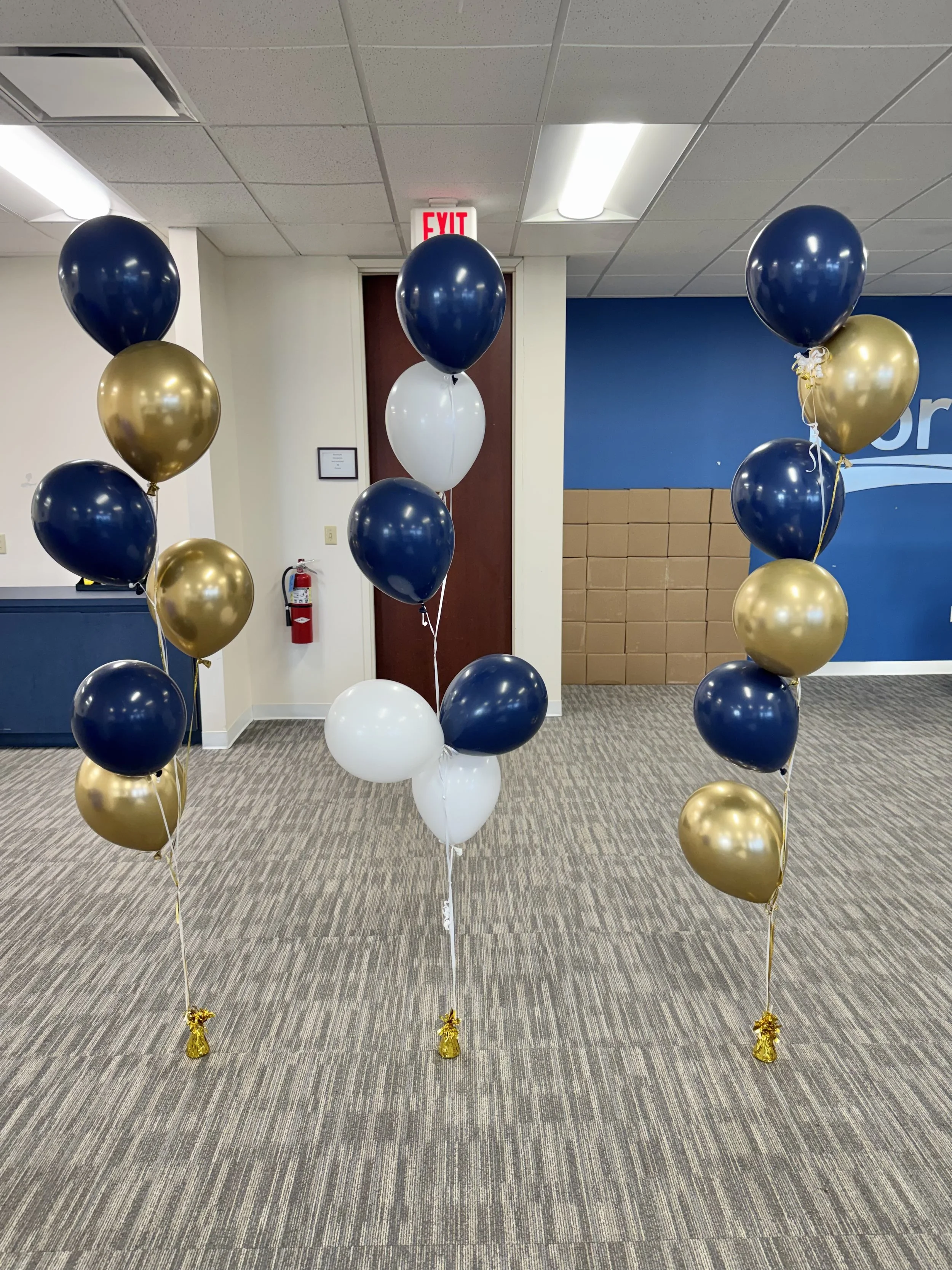 Three sets of helium balloons in navy, gold, and white tied to gold weights on a carpeted office or waiting room floor with a blue and beige wall in the background.