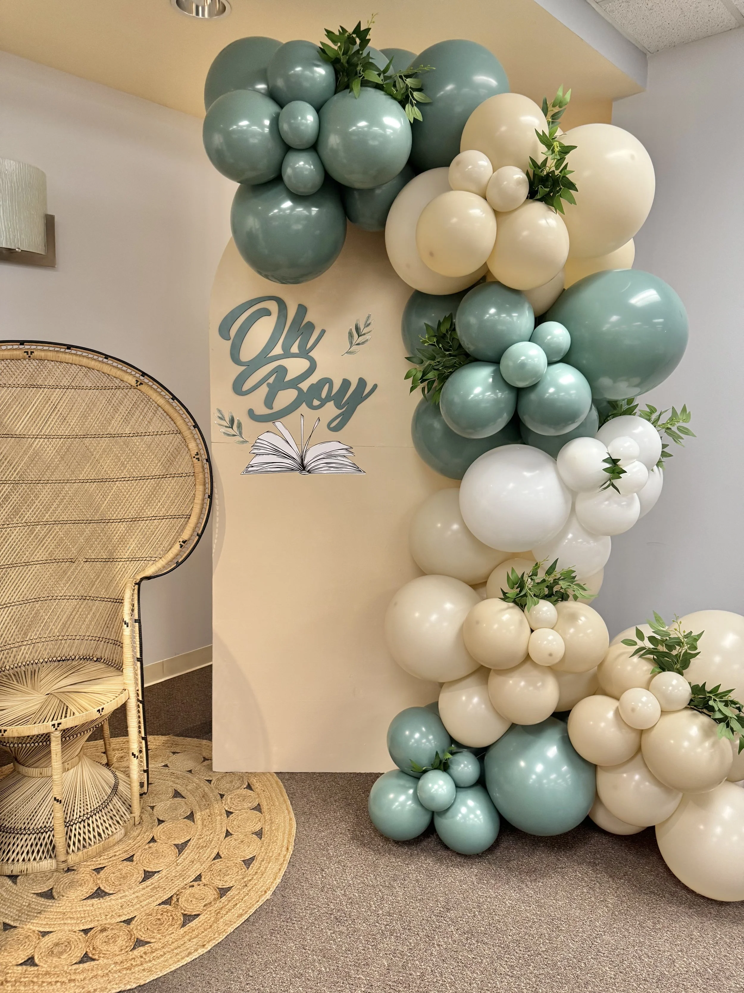 Decorative balloon display with green, white, and cream balloons arranged in a cascading pattern, accented with green leaves, next to a sign that reads "Oh Boy" with an open book illustration. A rattan chair and woven rug are visible nearby.