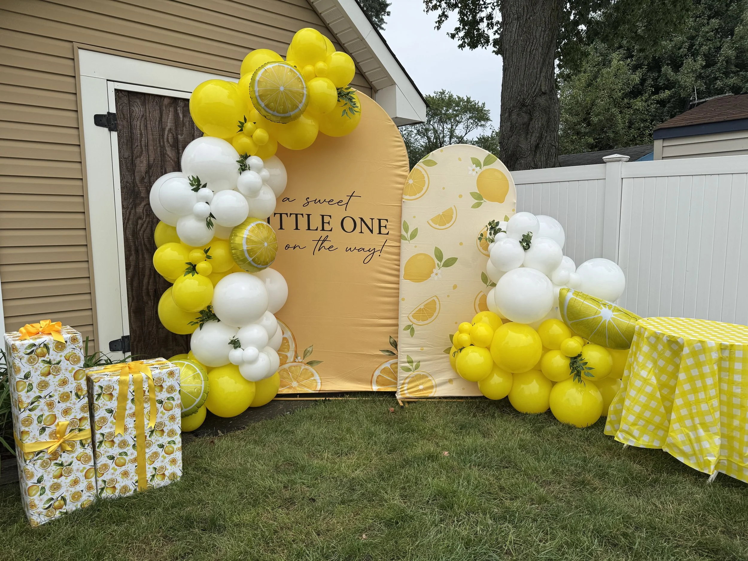 Decorations with yellow and white balloons, a large lemon-themed backdrop with the text "a sweet LITTLE ONE on the way!", wrapped lemon-patterned gifts, and a table covered with a yellow checkered tablecloth for a baby shower celebration.