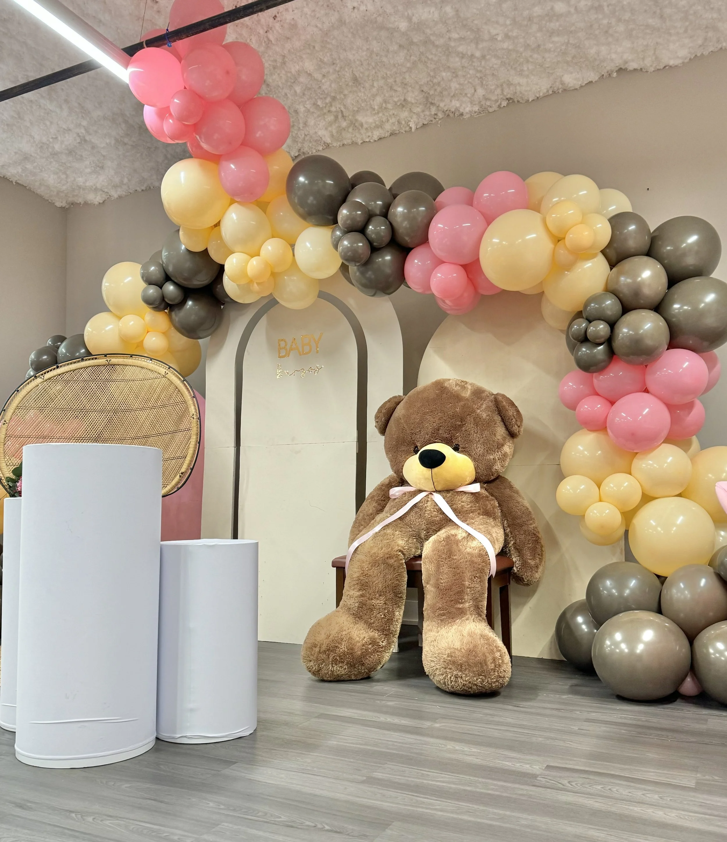Decorations for a baby shower with a large teddy bear, pastel balloon arch, and a white backdrop with the word 'BABY' on it.