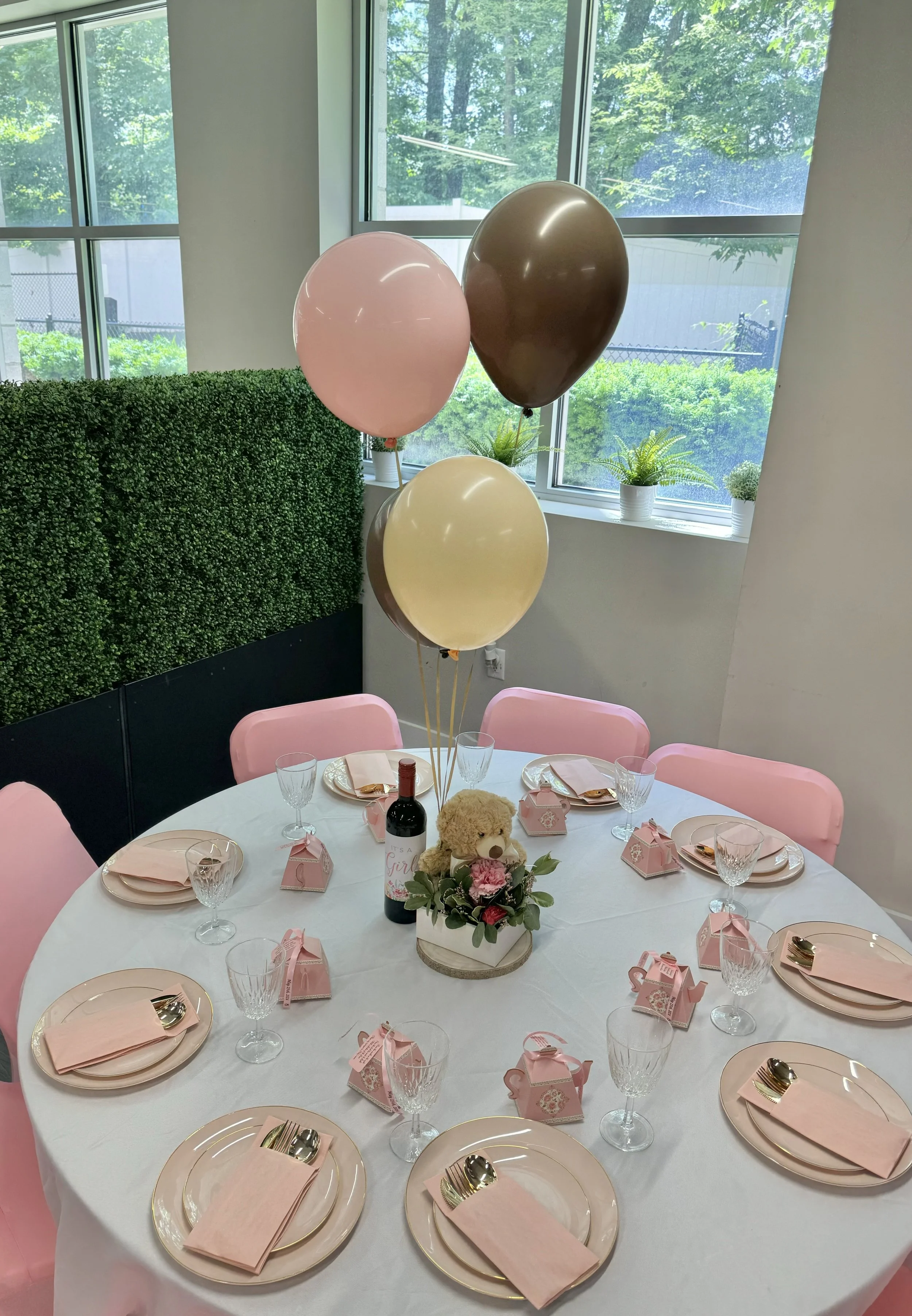 Decorated round table set for a celebration with pink and cream plates, pink napkins with gold utensils, pink chairs, floral centerpiece with a teddy bear, pink and gold balloons, and small pink gift boxes, in a room with large windows and green outd