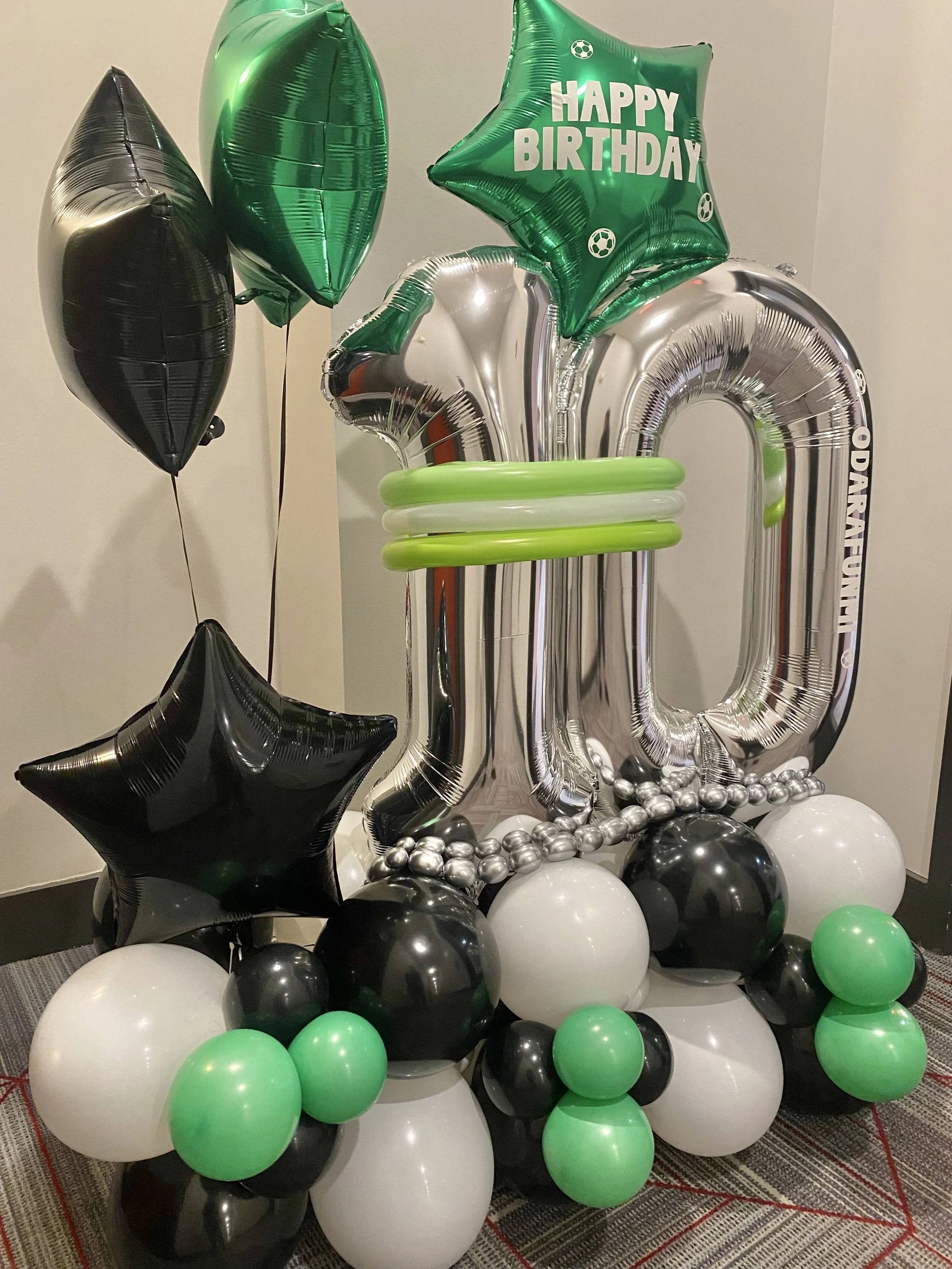 Celebration balloon arrangement with black, white, green, and silver balloons, including star-shaped and letter-shaped balloons, with a large silver number 10 balloon and a green star-shaped balloon that says "Happy Birthday."