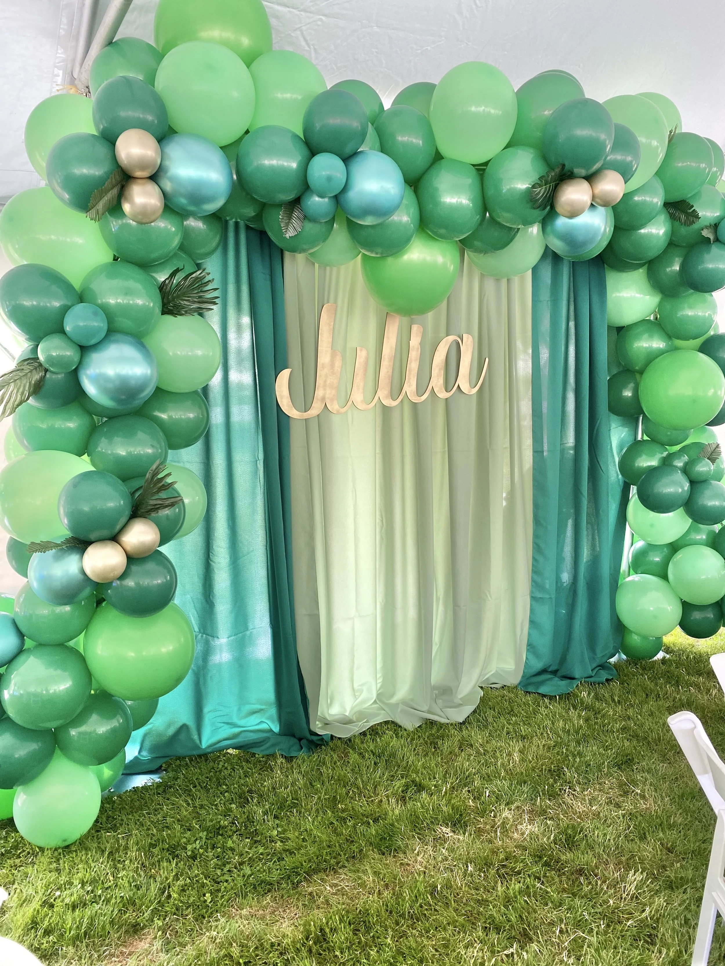 Decoration with green balloons, satin curtains, and a wooden sign reading 'Julia', likely at a celebration or party.