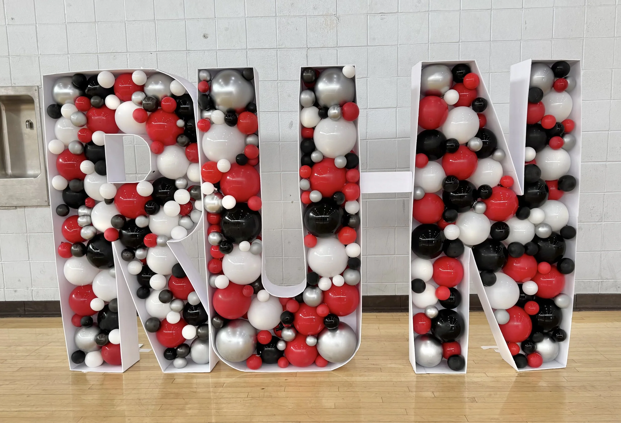 Large decorative letter 'R' filled with black, white, red, and silver balloons, placed against a white tiled wall.