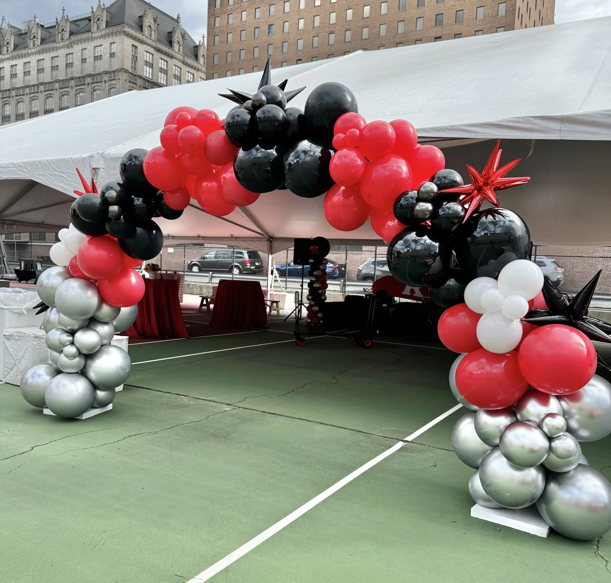 Balloon arch made of red, black, white, and silver balloons, with star-shaped balloons on top, set up under a white event tent in an outdoor urban area.