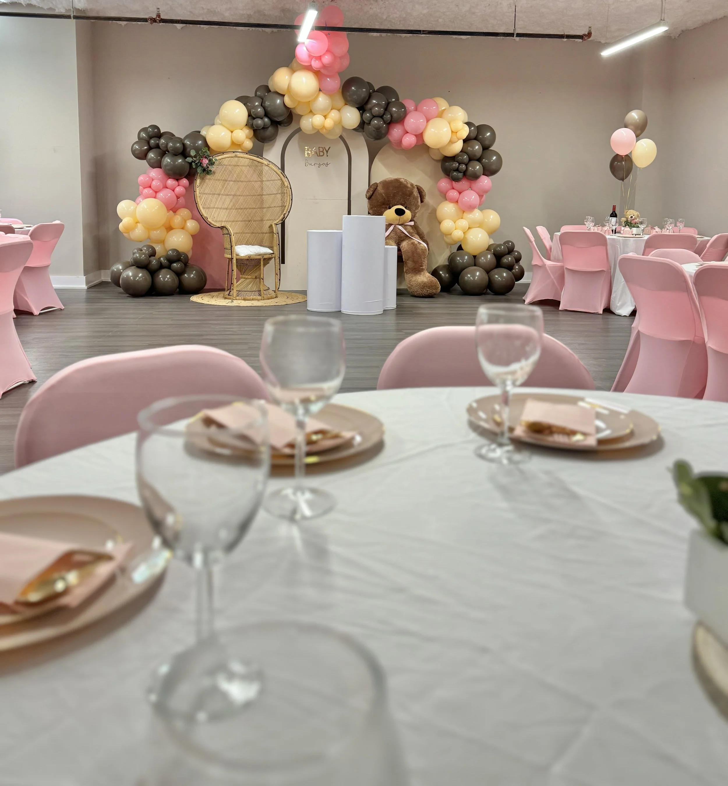 Baby shower decorations featuring pink, beige, and gray balloons forming an arch, with a teddy bear, decorative chairs, and a table set for guests.