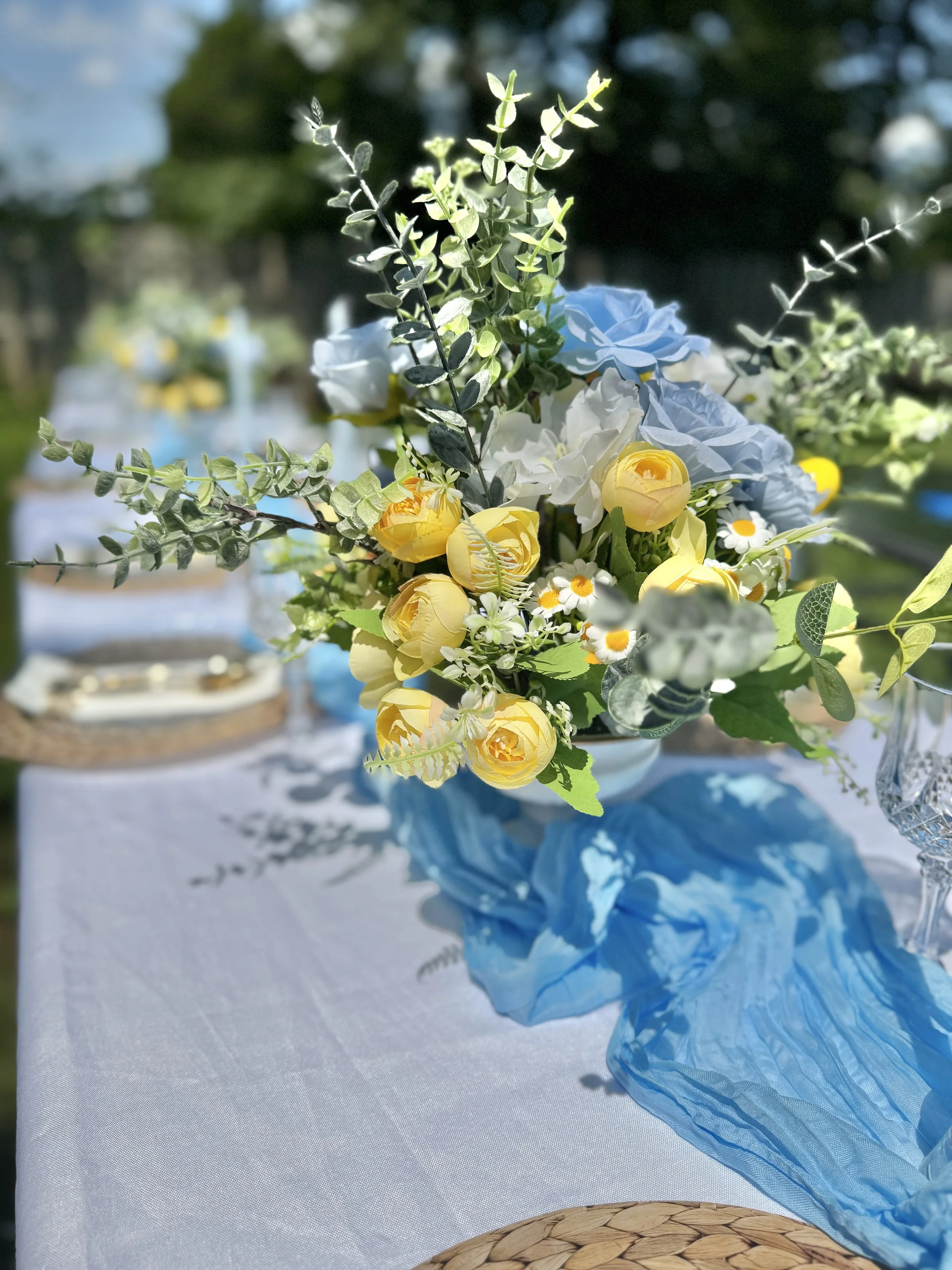 A close-up of a floral centerpiece with blue, yellow, and white flowers and green foliage, set on a table with a white tablecloth and a blue fabric runner. There are blurred outdoor trees in the background.