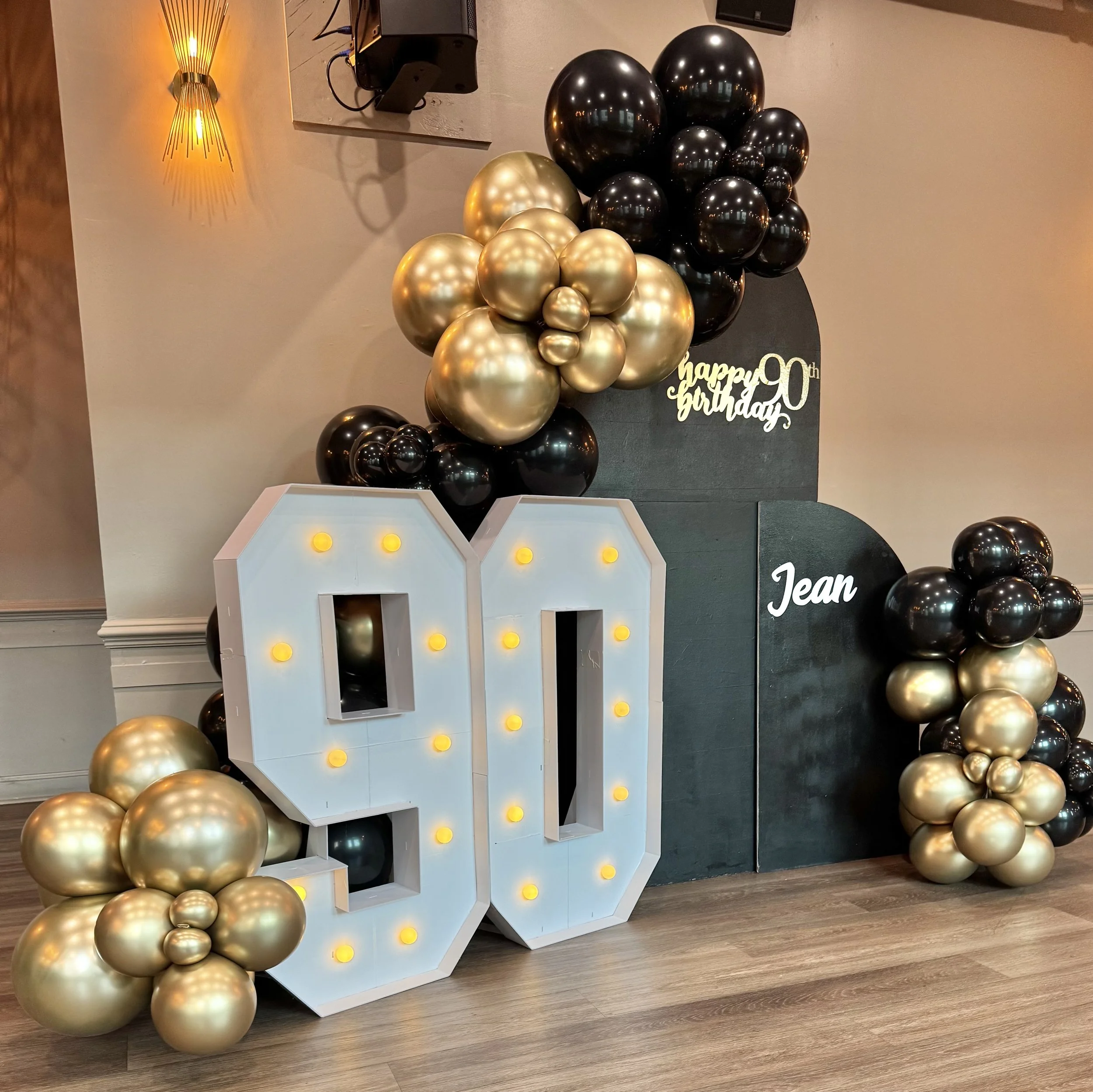 Gold and black balloons arranged around a white decorative letter 'J' with yellow lights, celebrating a 20th birthday.