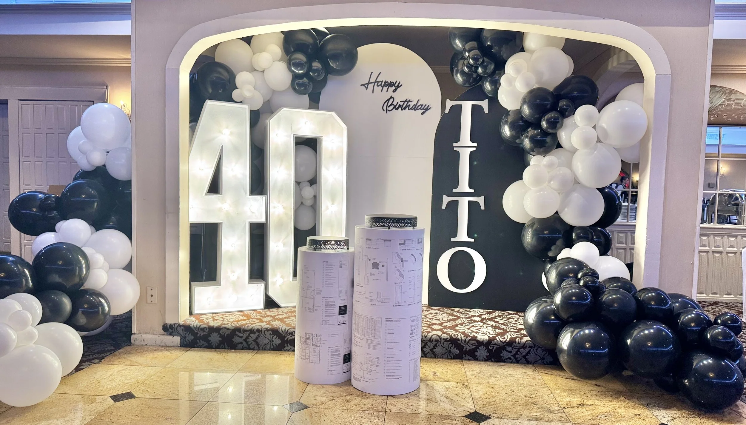 Decorative birthday display for a 40th celebration with a large illuminated '40', white and black balloons, and a backdrop with 'Happy Birthday TITio' written on it.