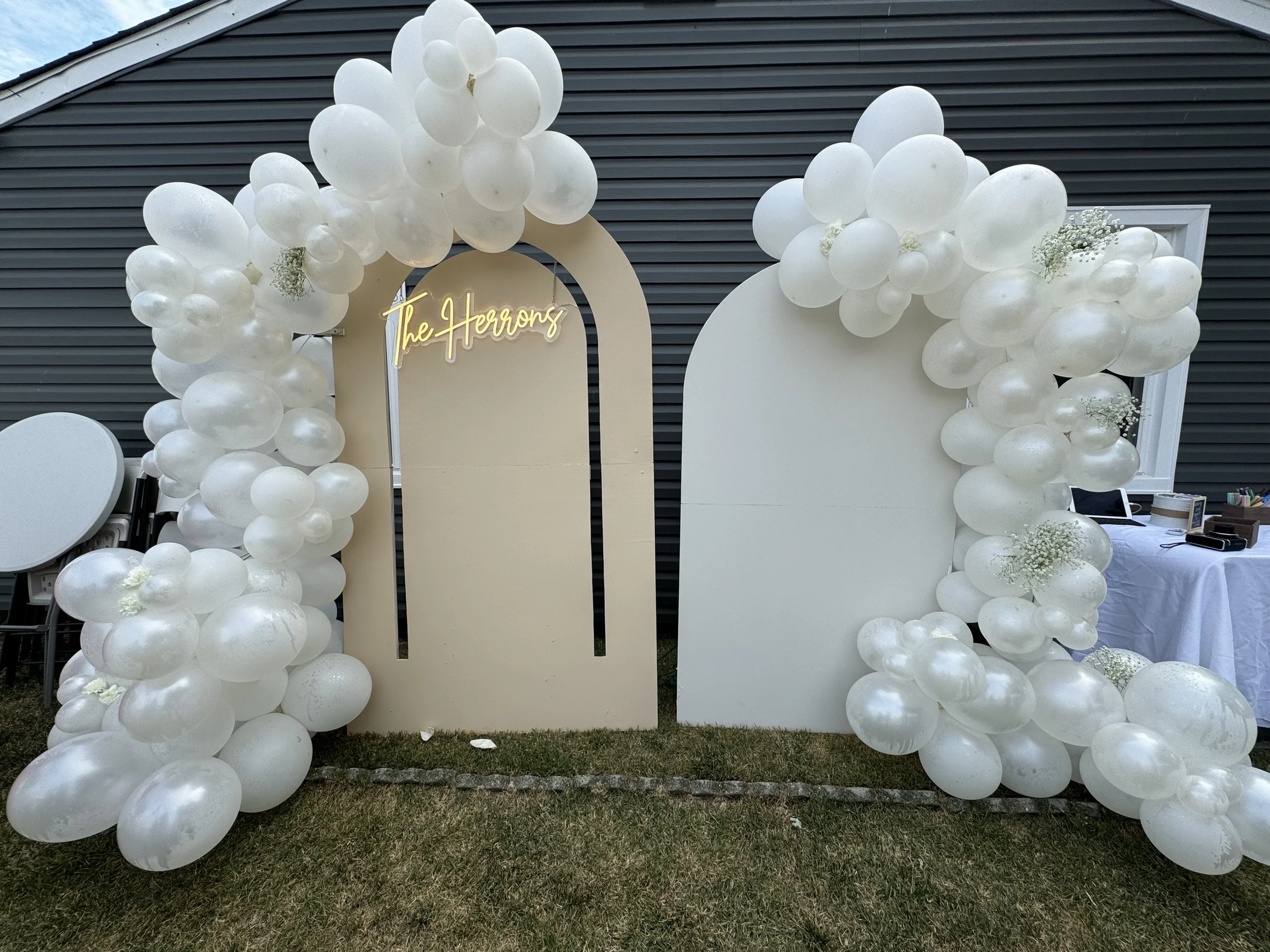 Decorative event backdrop with white balloons, white panels, a neon sign reading 'The Harrons,' and a table with items on the right side.
