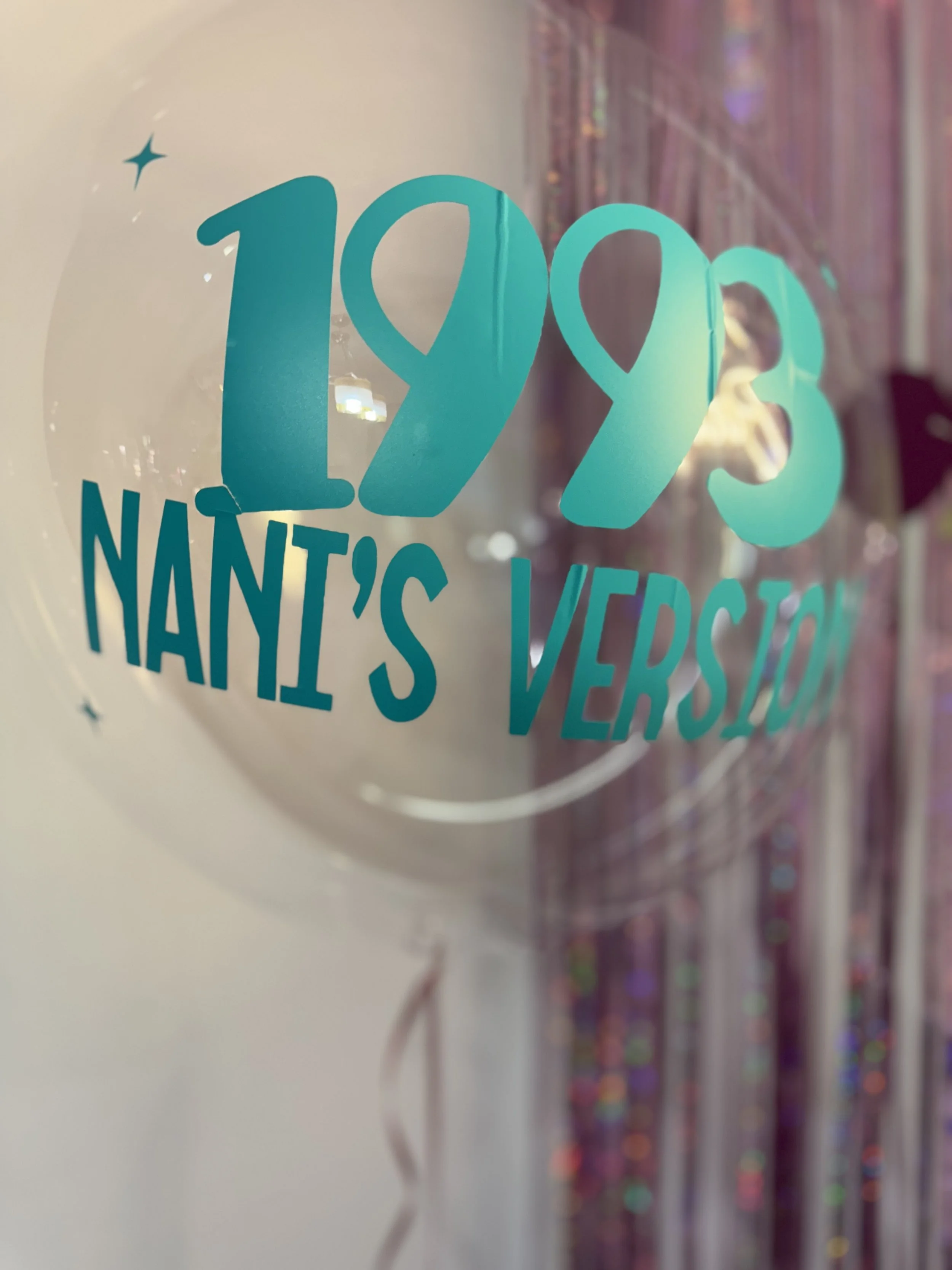 A transparent balloon with teal lettering reading '1993 NANI'S VERSION' and small star designs, reflecting a celebration or anniversary.