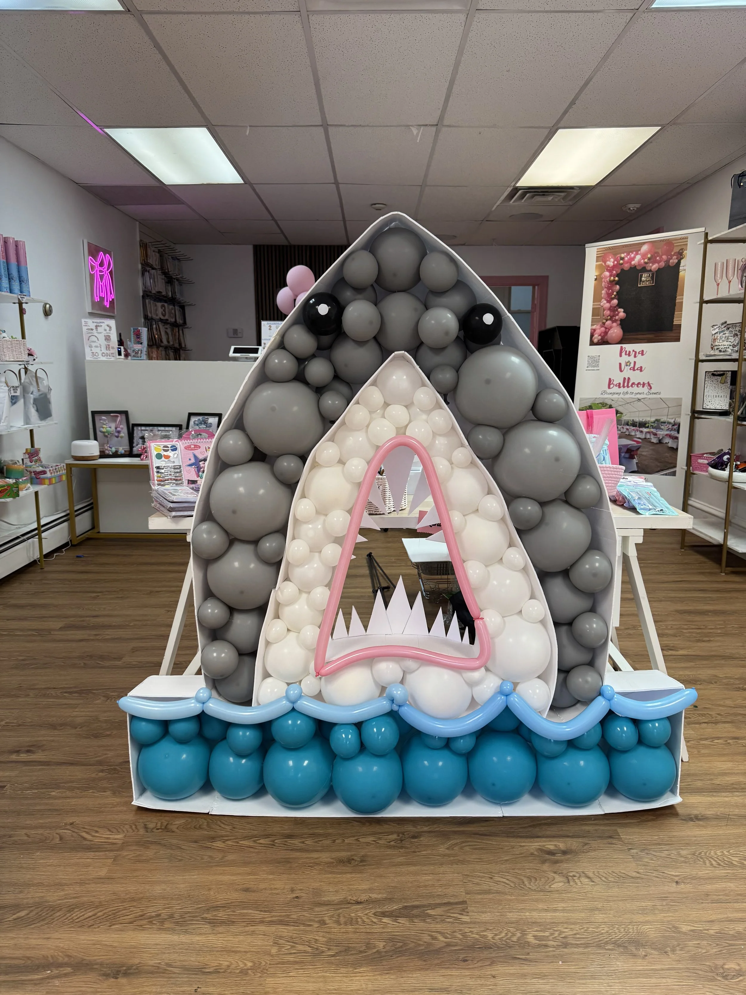 Balloon display shaped like a shark with a pink balloon outline of sharp teeth, set on a white base with blue balloons at the bottom, in a store with balloons and decorative items.