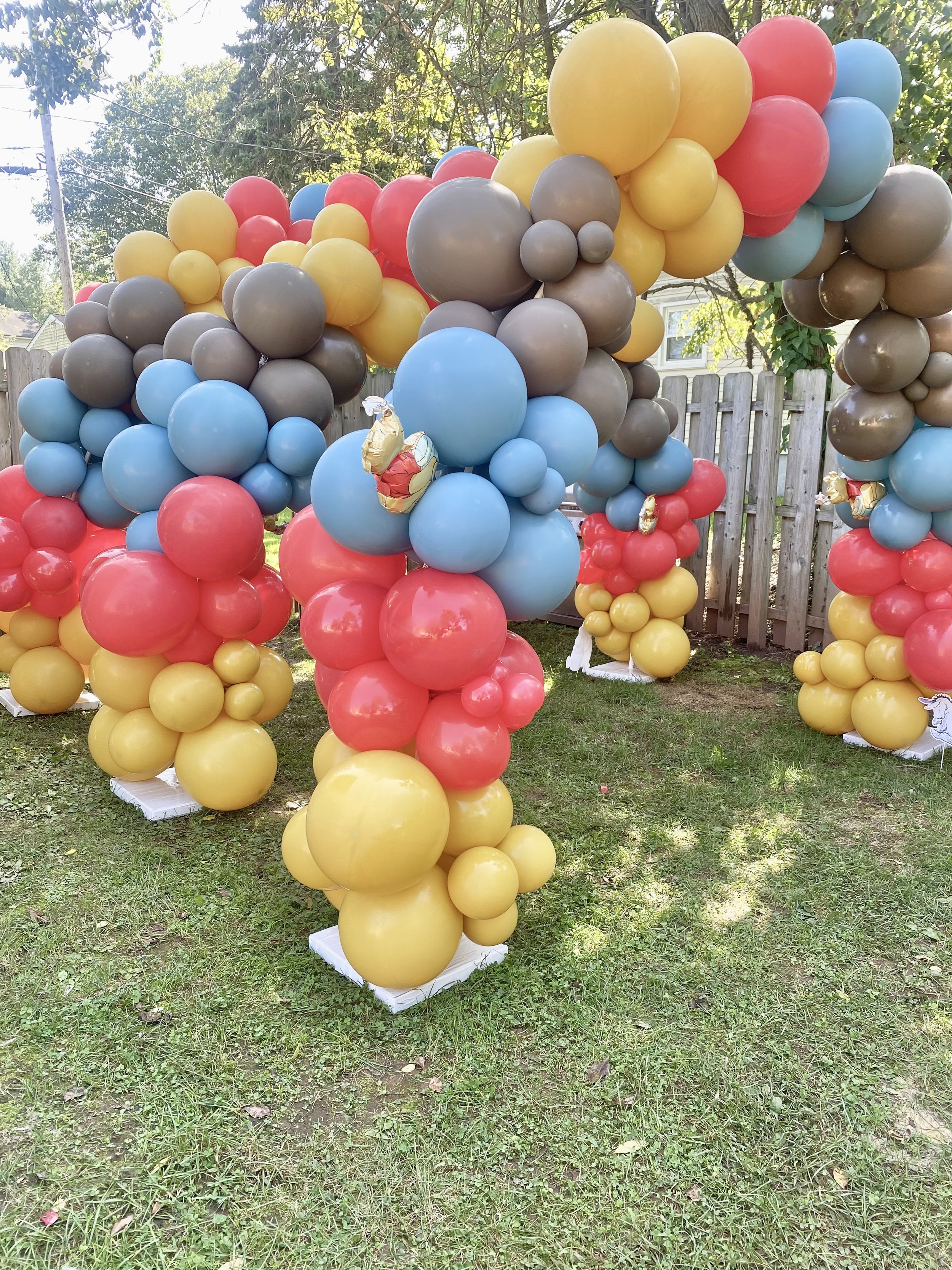 Colorful balloon arrangement with red, yellow, blue, and gray balloons, decorated with small shiny balloon animals, set outdoors on grass with a wooden fence and trees in the background.