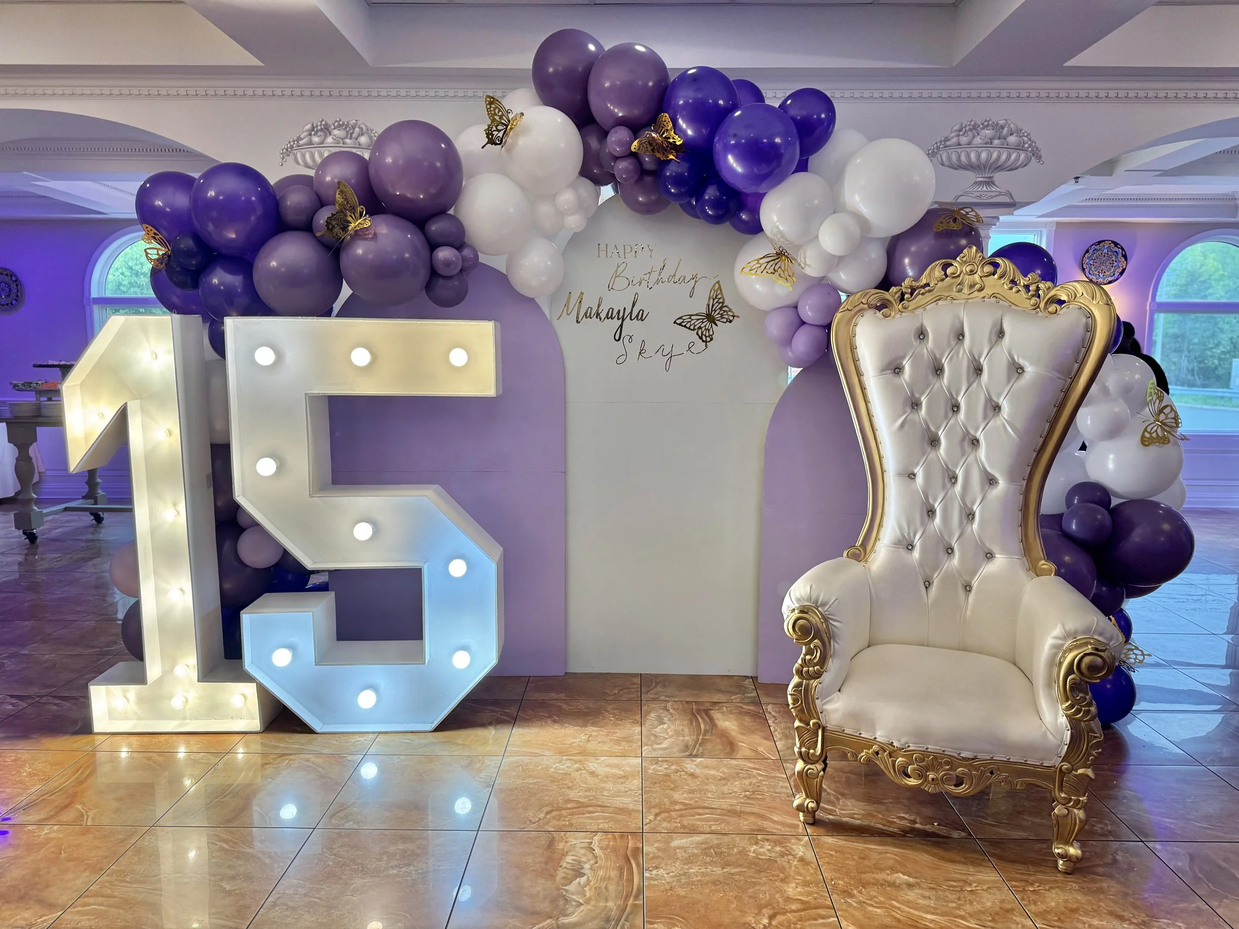Decorative birthday setup with large illuminated number 15, purple and white balloons arch, gold-trimmed white throne, and a sign with 'Happy Birthday Makayla Sky' in a room with polished marble floor and large windows.