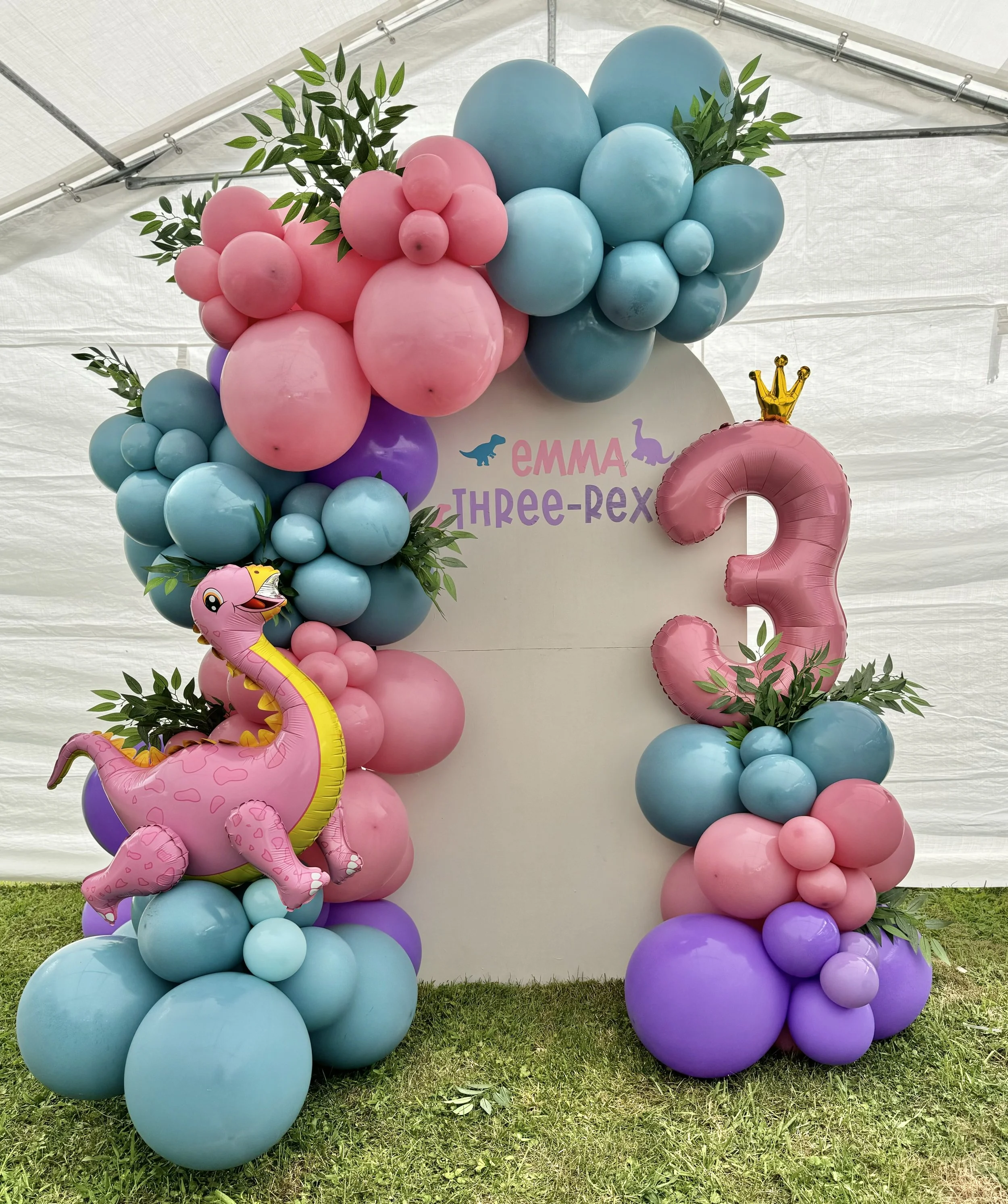 Party decor with pastel balloons in pink, blue, purple, and green arranged around a white backdrop with the name Emma and the title Three-Rex, along with dinosaur-themed decorations and a pink number three balloon with a gold crown.