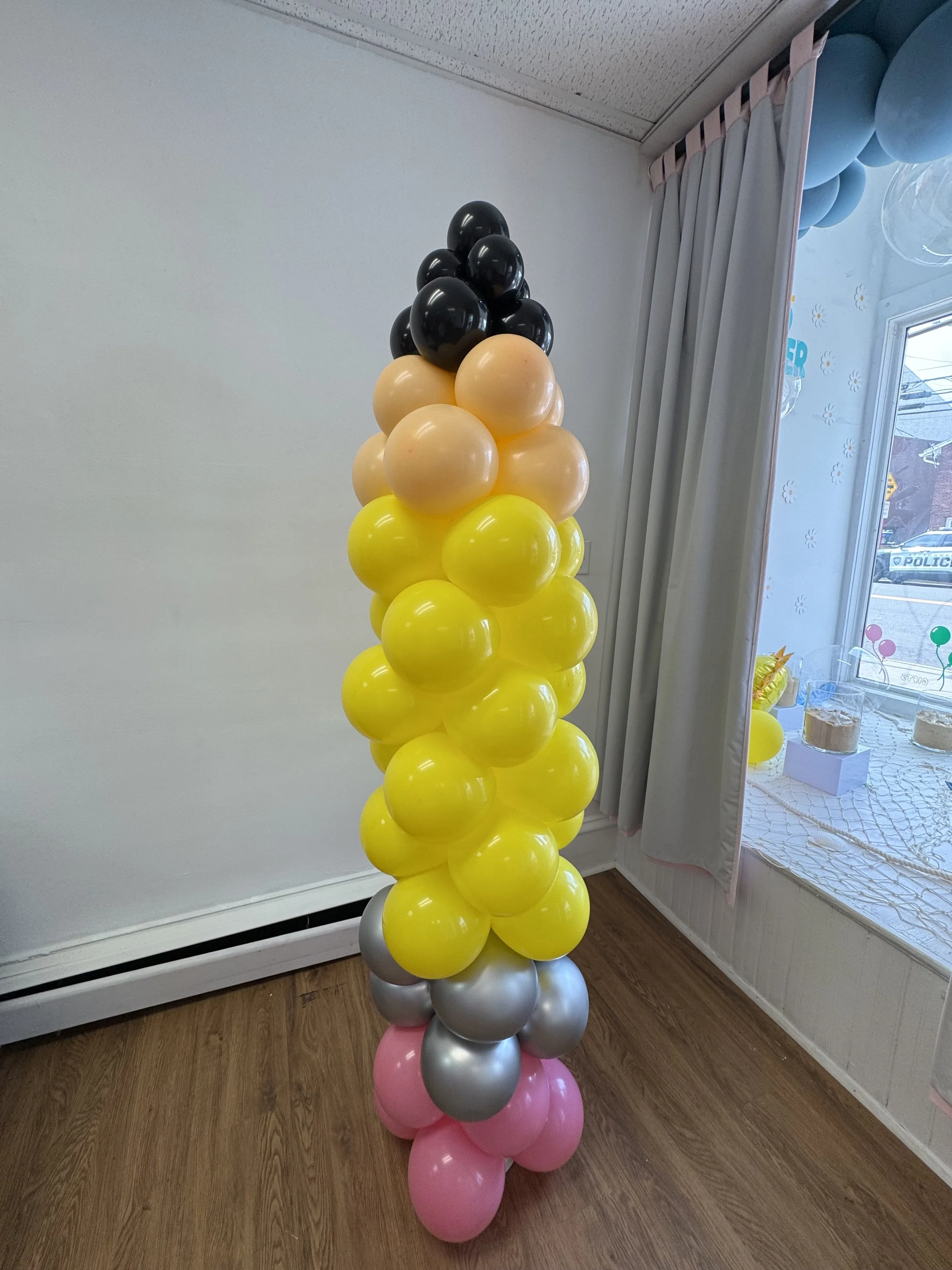 A tall column of balloons arranged in color sections: pink at the bottom, followed by silver, yellow, peach, black, and pink again at the top, standing on a wooden floor inside a room near a window with curtains.